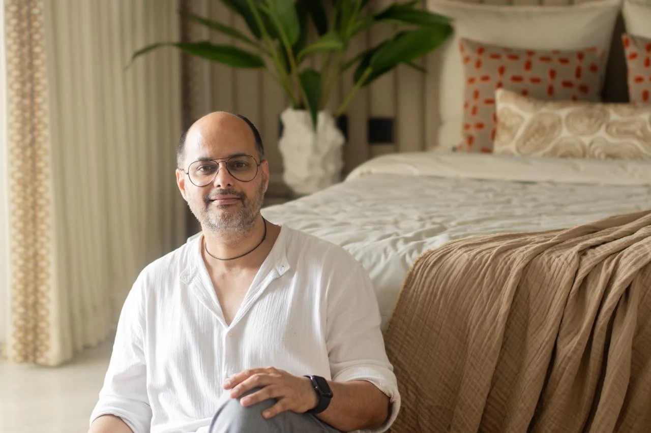 A man with glasses, a beard, and wearing a white shirt, sitting in a bedroom with a large bed, a beige blanket, multiple pillows, a large plant, and curtains in the background.