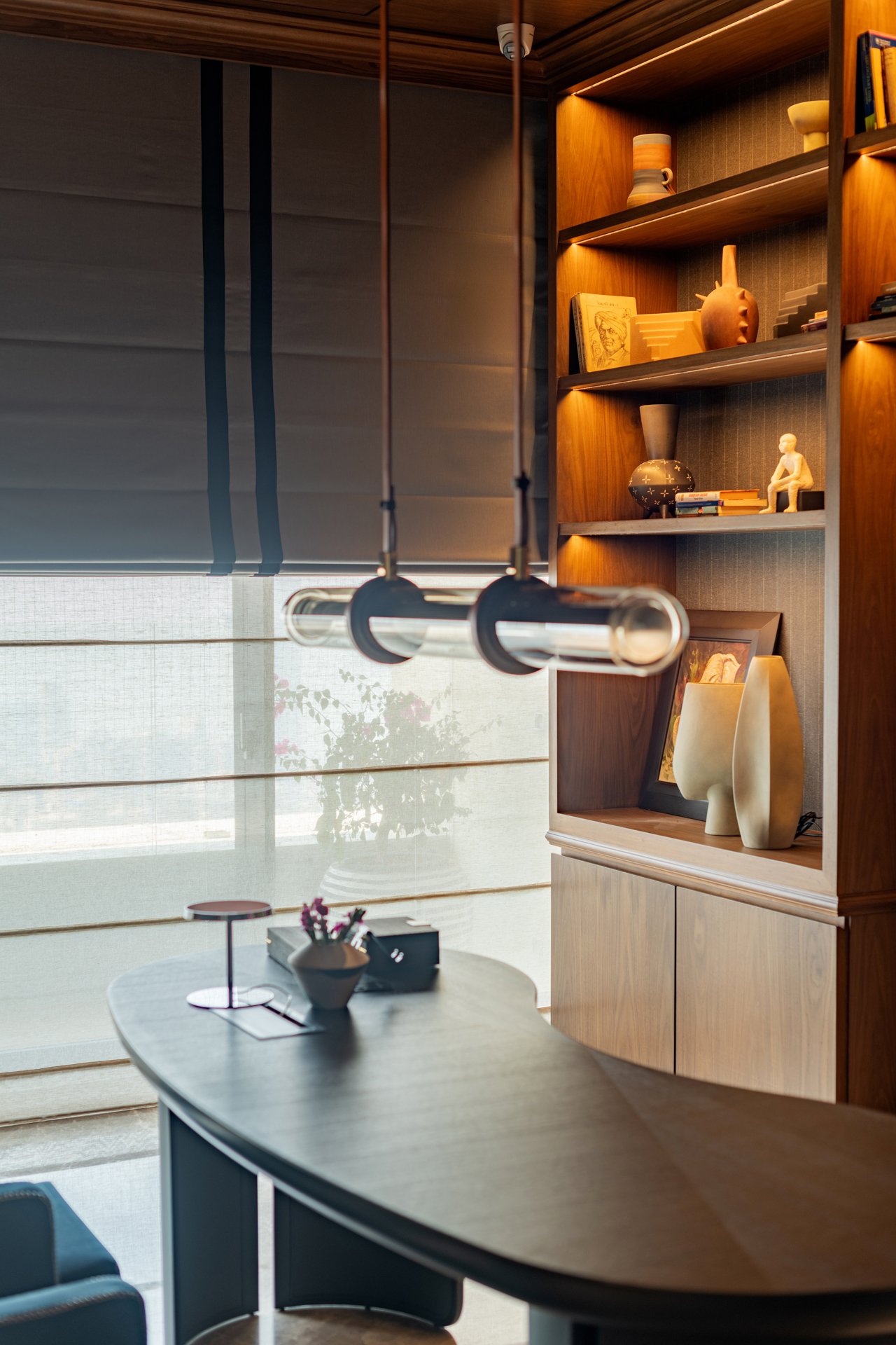 Interior view of a modern room with a wooden bookshelf filled with decorative items and books, a table with a small lamp, a bowl with flowers, and a few office supplies, near a large window with blinds.
