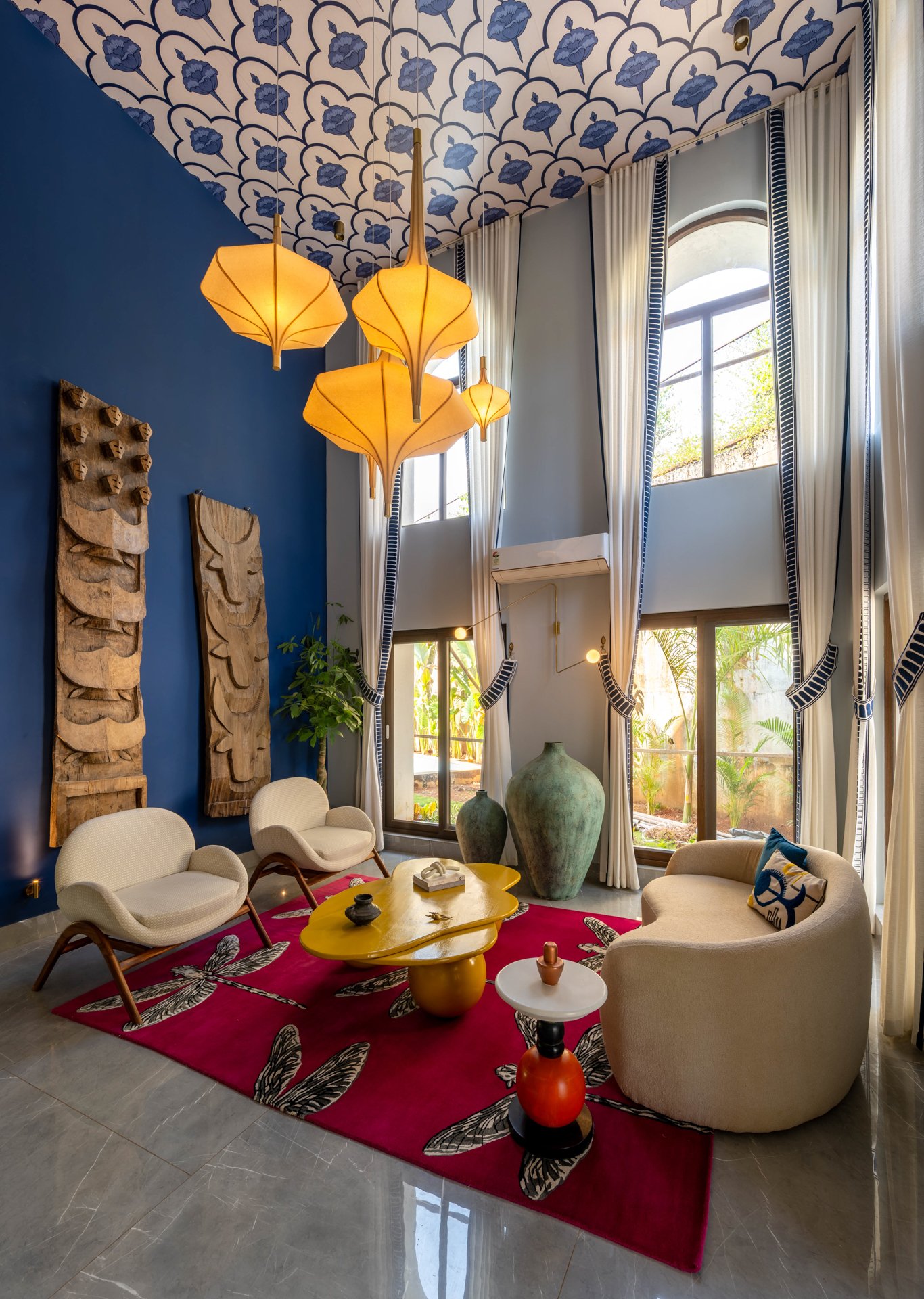 A living room with high ceilings featuring a patterned ceiling design and large windows with white curtains. There are modern white chairs, a yellow coffee table, and a red rug with leaf patterns. The walls are decorated with wooden tribal art pieces and potted plants. Hanging yellow pendant lights illuminate the space.