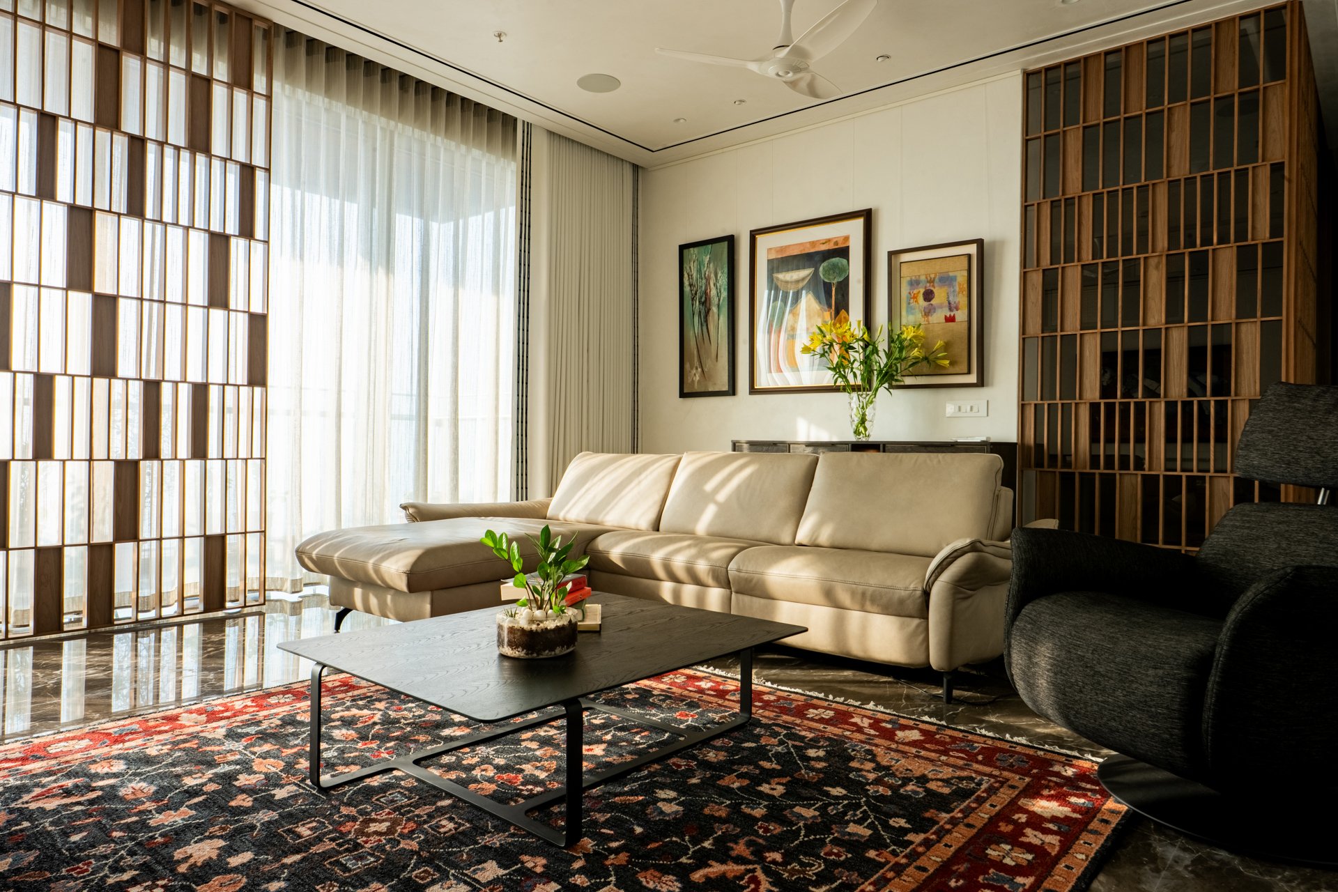 Living room with beige sofa, black armchair, wooden room divider, artwork on the wall, vase with yellow flowers, coffee table with plant, patterned area rug, large window with sheer curtains, ceiling fan.
