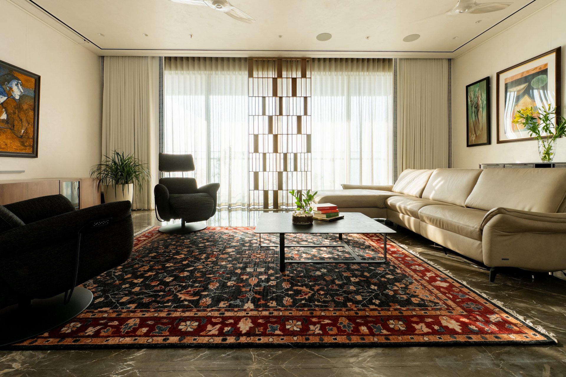 Living room with a beige sectional sofa, black and gray chairs, a marble coffee table with a small plant and books, Persian rug, wall art, plants, and large windows with curtains.