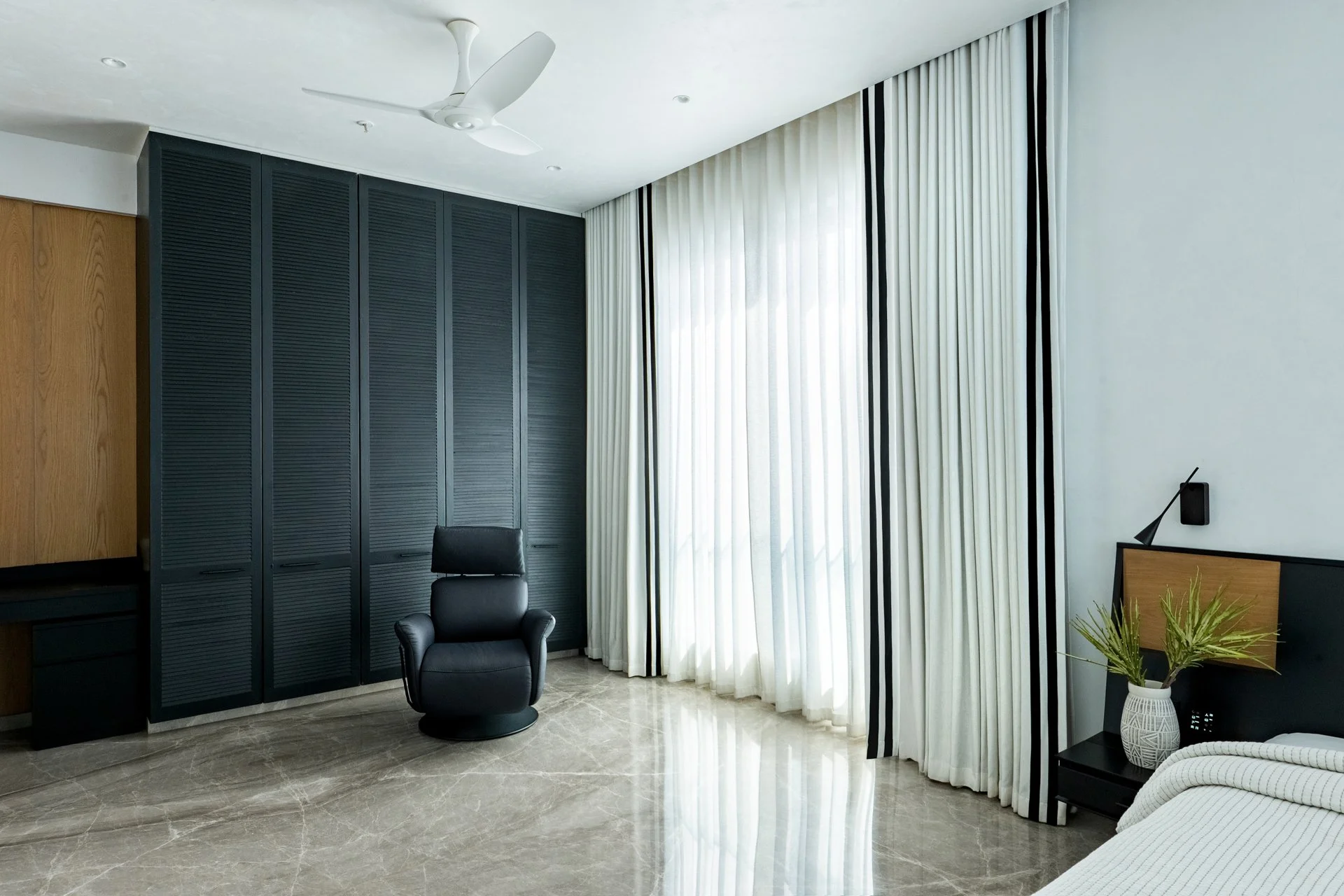 Modern living room with black leather armchair, tall black cabinet, sheer curtains, white ceiling fan, potted plant in a white vase, marble floor, and part of a white sofa.