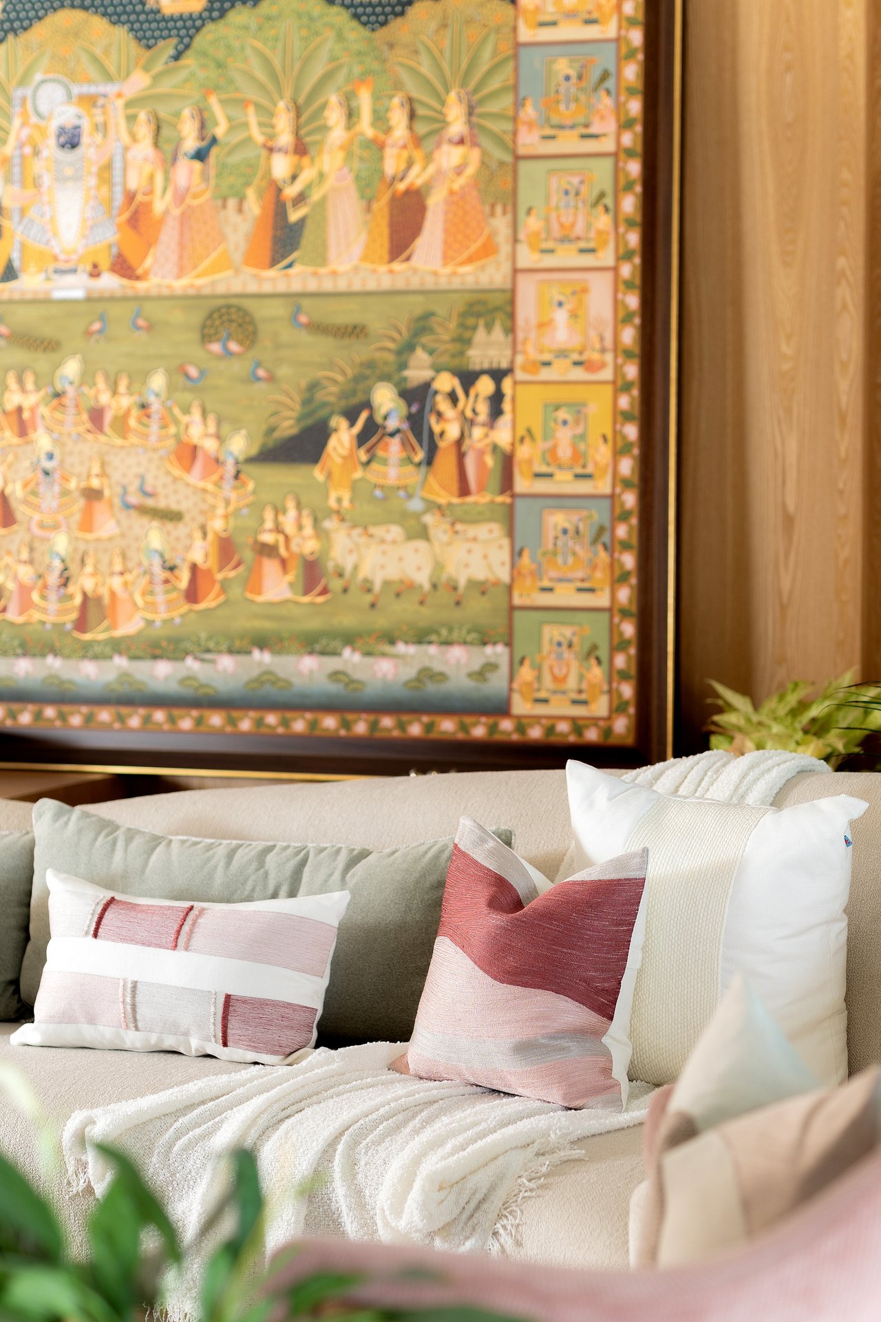 Close-up of a living room corner with a beige sofa, decorative pillows in pink, white, and beige, a white throw blanket, and a large colorful traditional painting on the wall featuring figures in a natural landscape.