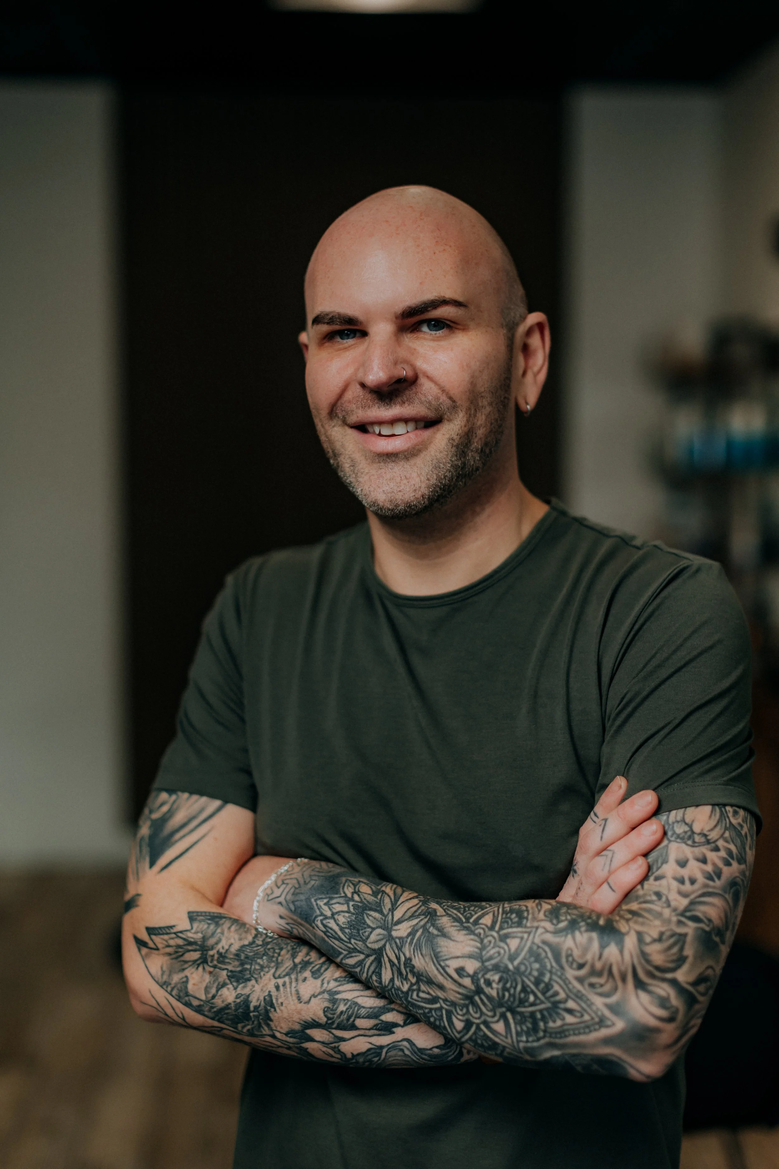 A smiling man with tattoos on his arms, a nose piercing, and a shaved head, wearing a dark green T-shirt, standing indoors with arms crossed.
