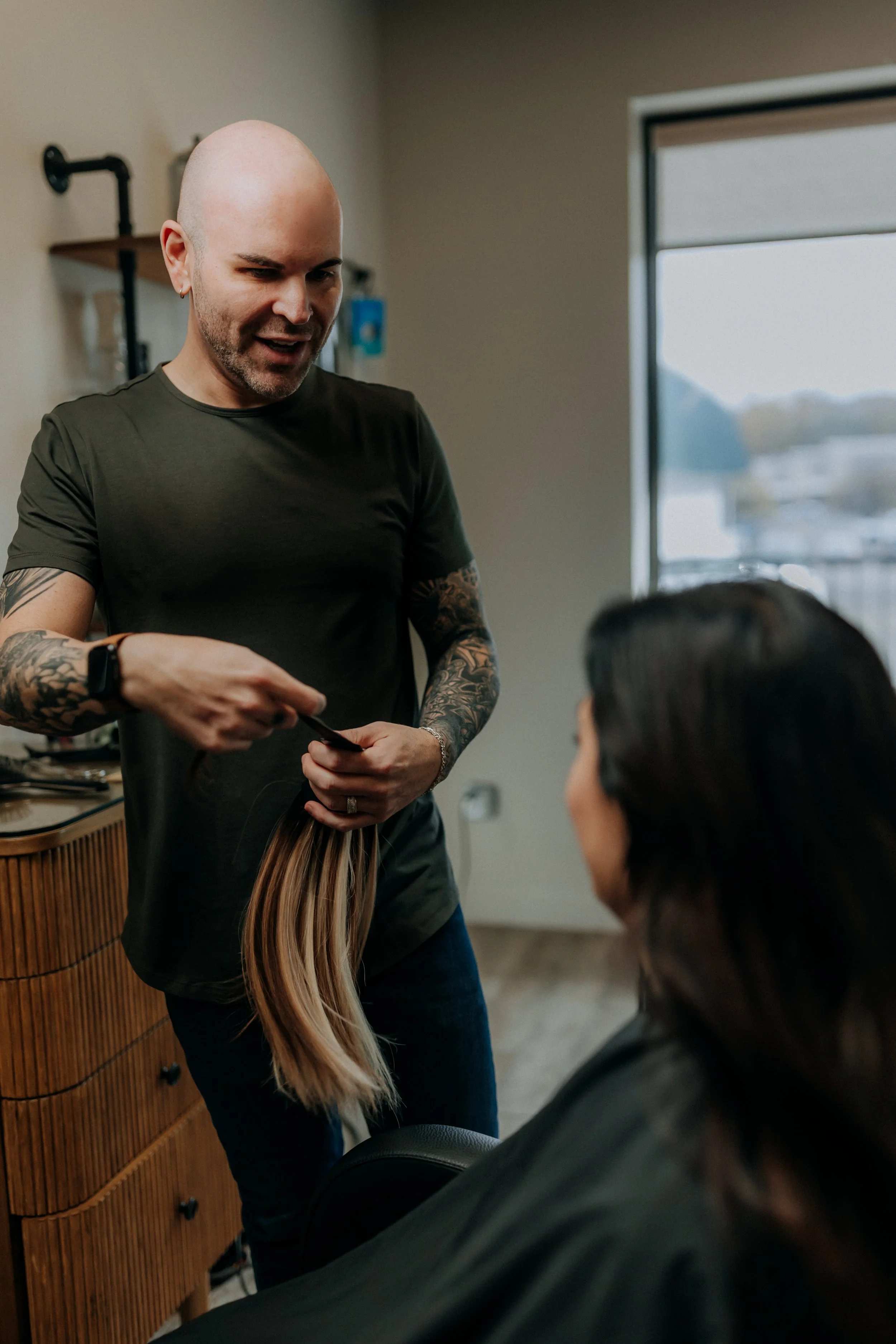 A male hairstylist with tattoos on his arms holding a hair extension and smiling at a woman sitting in a salon chair, inside a modern hair salon with a large window in the background.