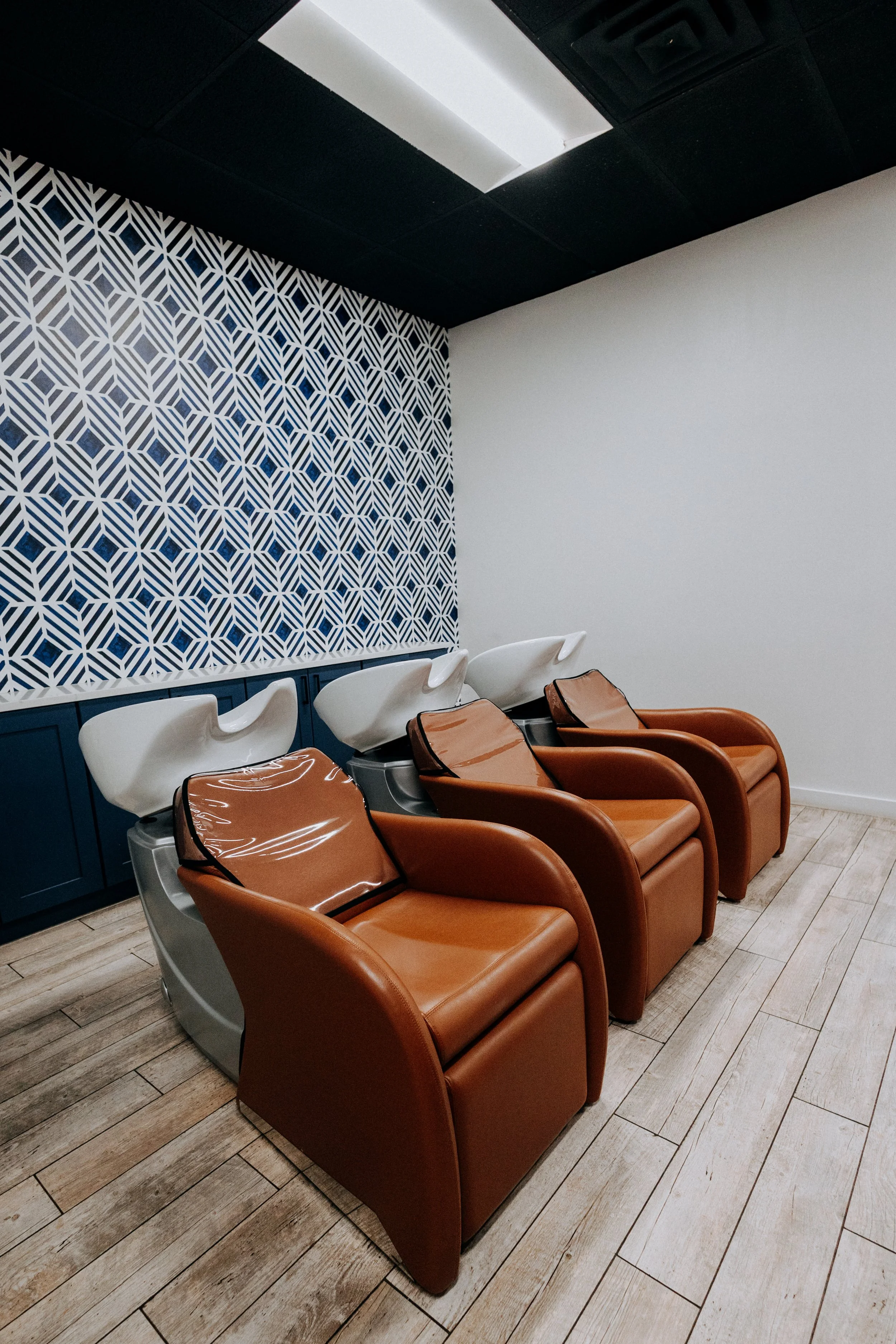 Three salon chairs with attached hair wash basins in a modern salon or spa, with a decorative geometric patterned wall and wooden flooring.