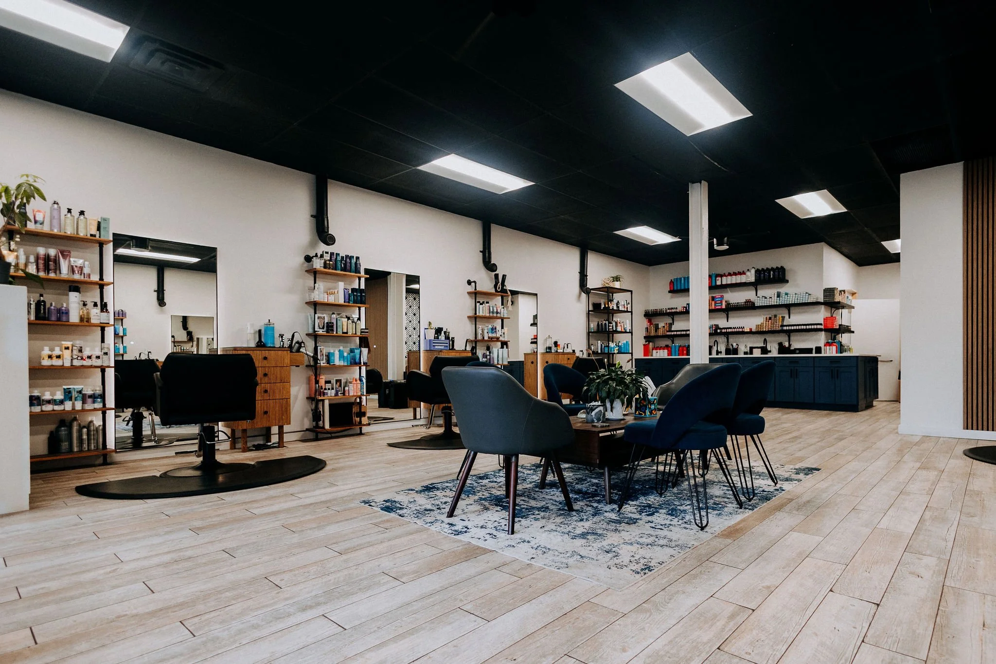 Interior of a modern hair salon with styling stations, chairs, mirrors, shelves stocked with hair products, a seating area with a table, plants, and bright overhead lighting.