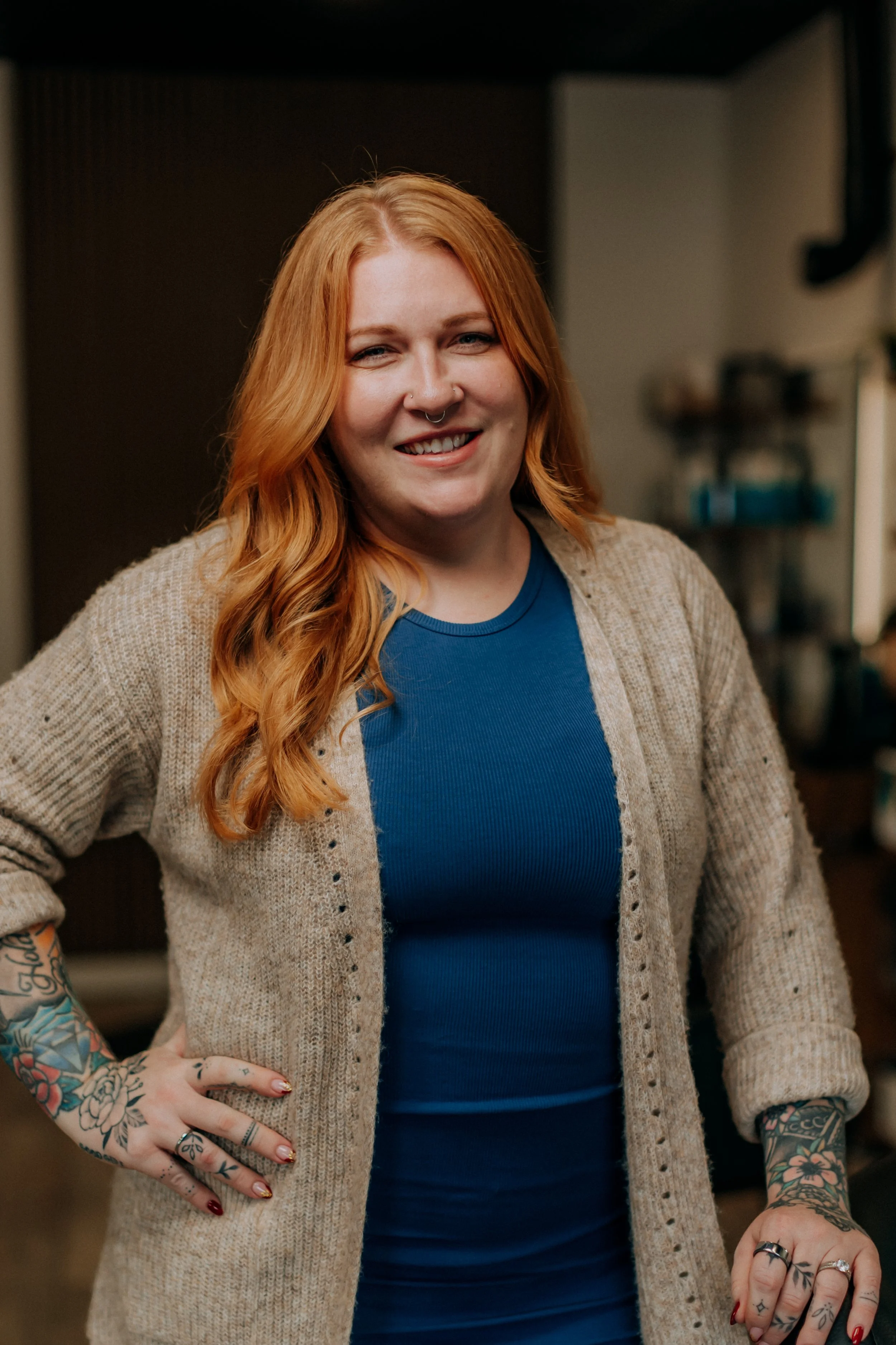 A woman with long wavy red hair, tattoos on her arms, and multiple rings on her fingers, smiling and standing indoors.