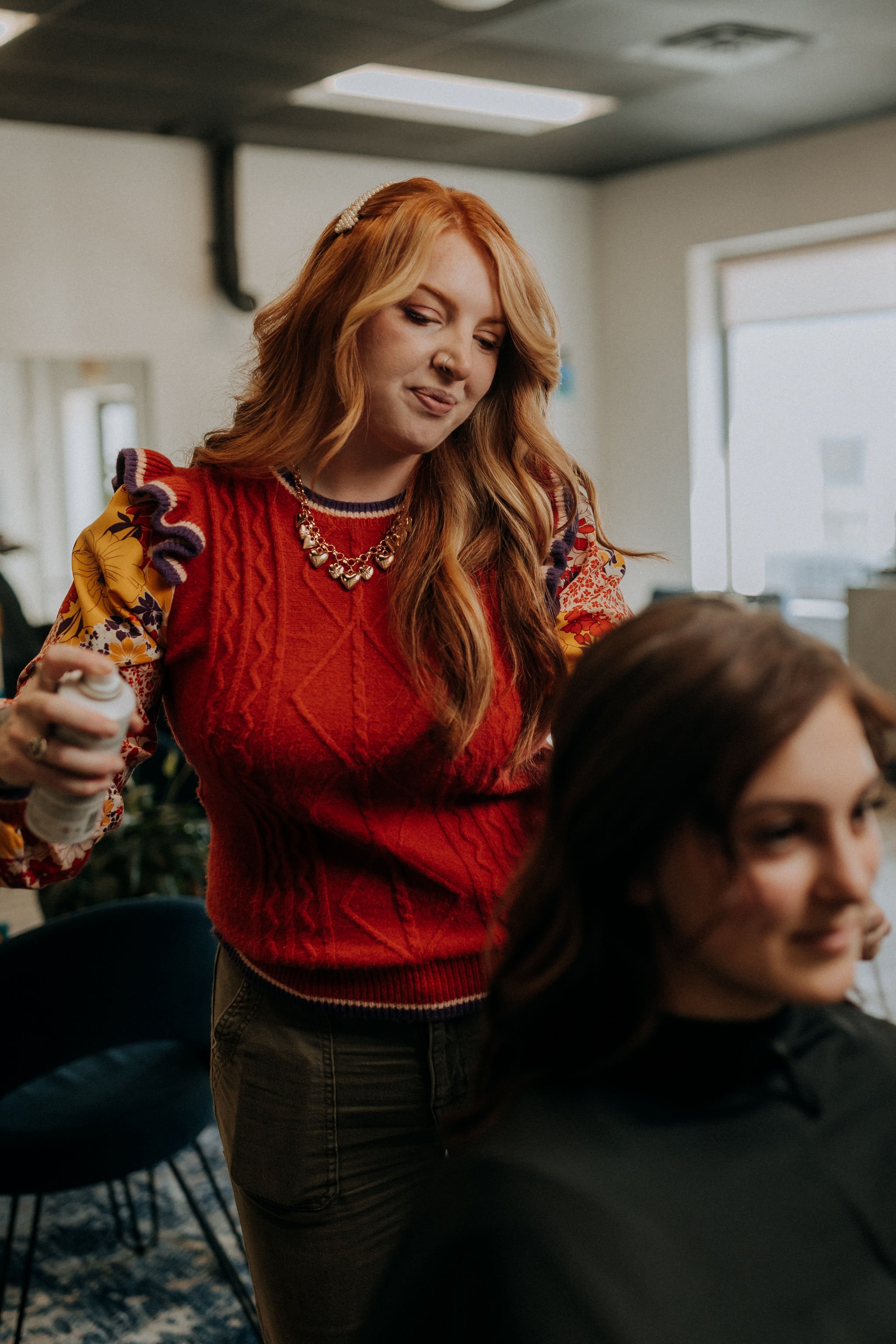 A woman with long red hair and a colorful, patterned sweater is standing behind another woman with dark brown hair, who is sitting in a salon chair.
