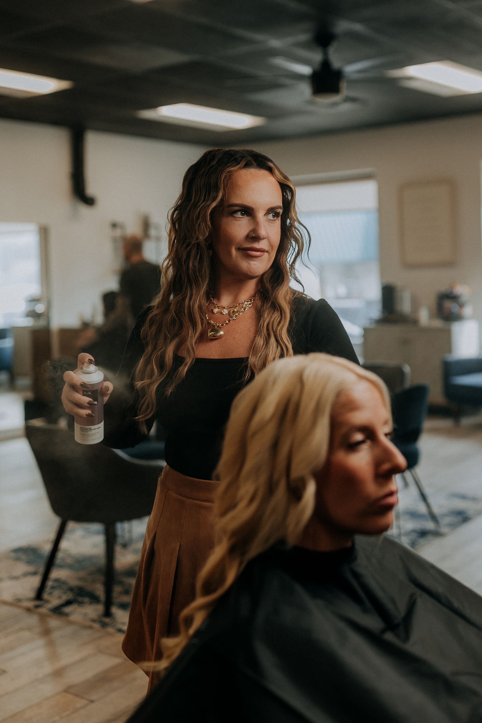 A woman with wavy brown hair and gold jewelry is standing in a salon, holding a spray can, while another woman with blonde hair sits with her eyes closed, wearing a black cape.