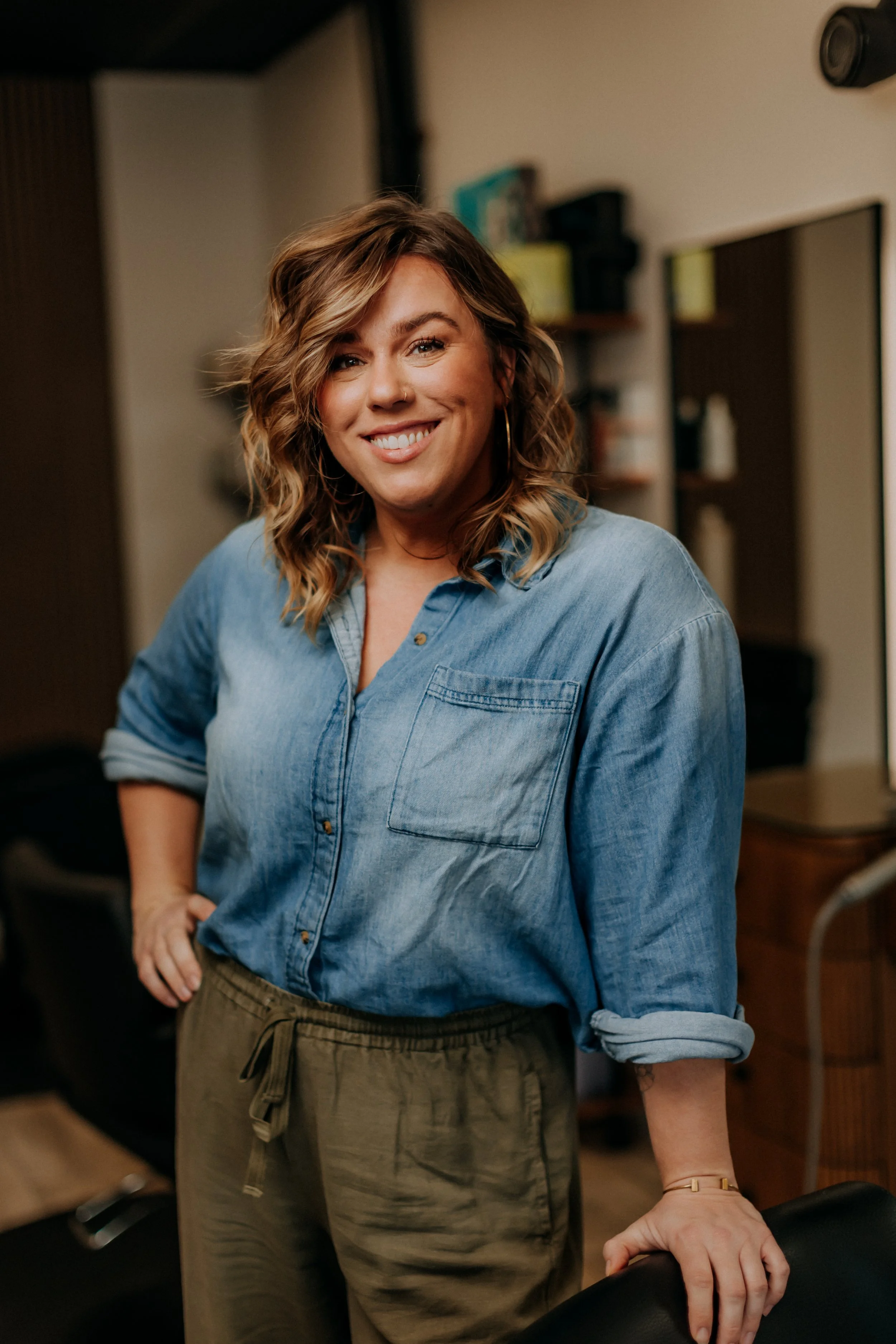 A woman with shoulder-length wavy hair smiling, wearing a denim button-down shirt with rolled-up sleeves and olive green pants, standing indoors in a casual setting.