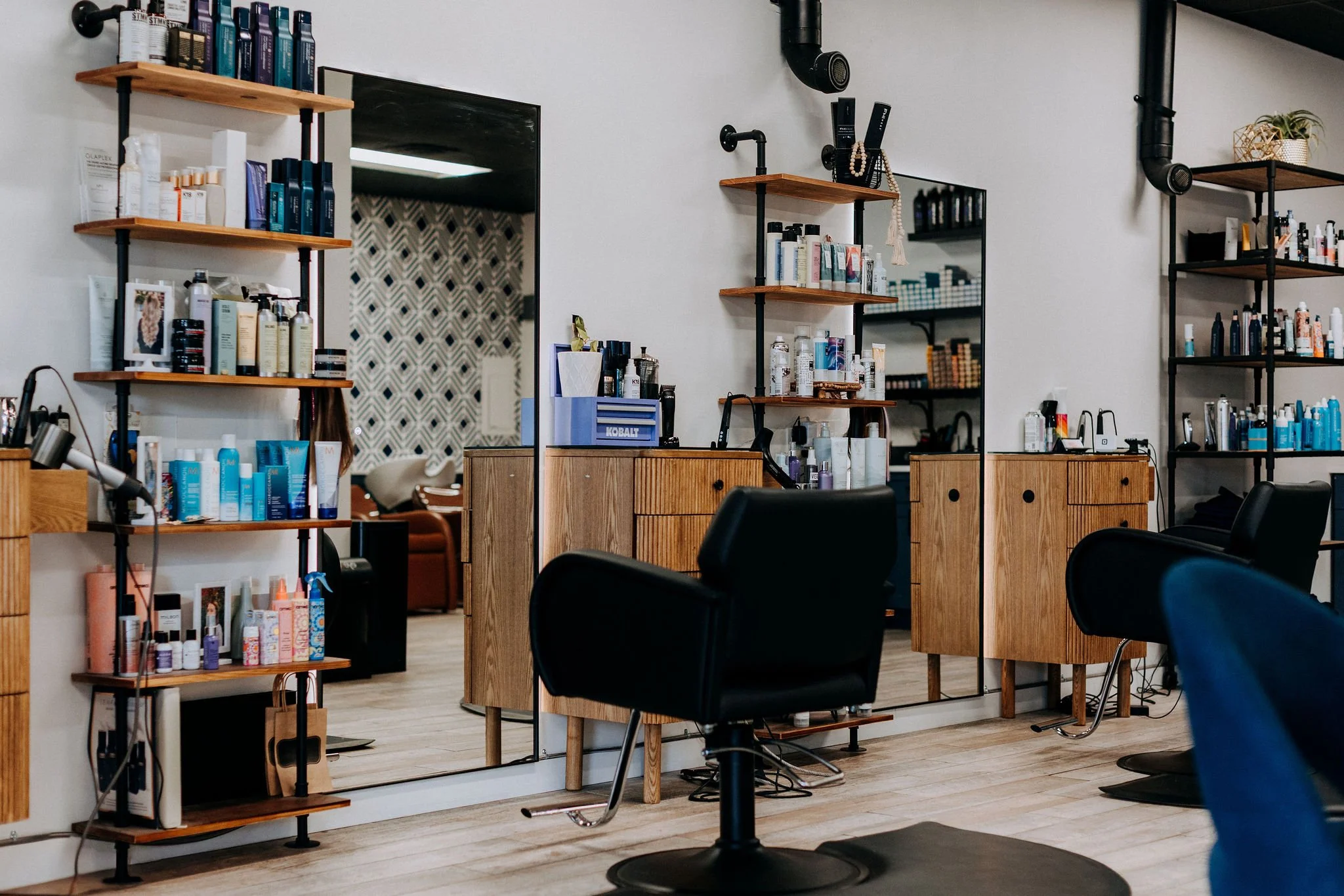 Interior of a hair salon or barber shop with black chairs, wooden stations, shelves with hair products, and a large mirror on the wall.