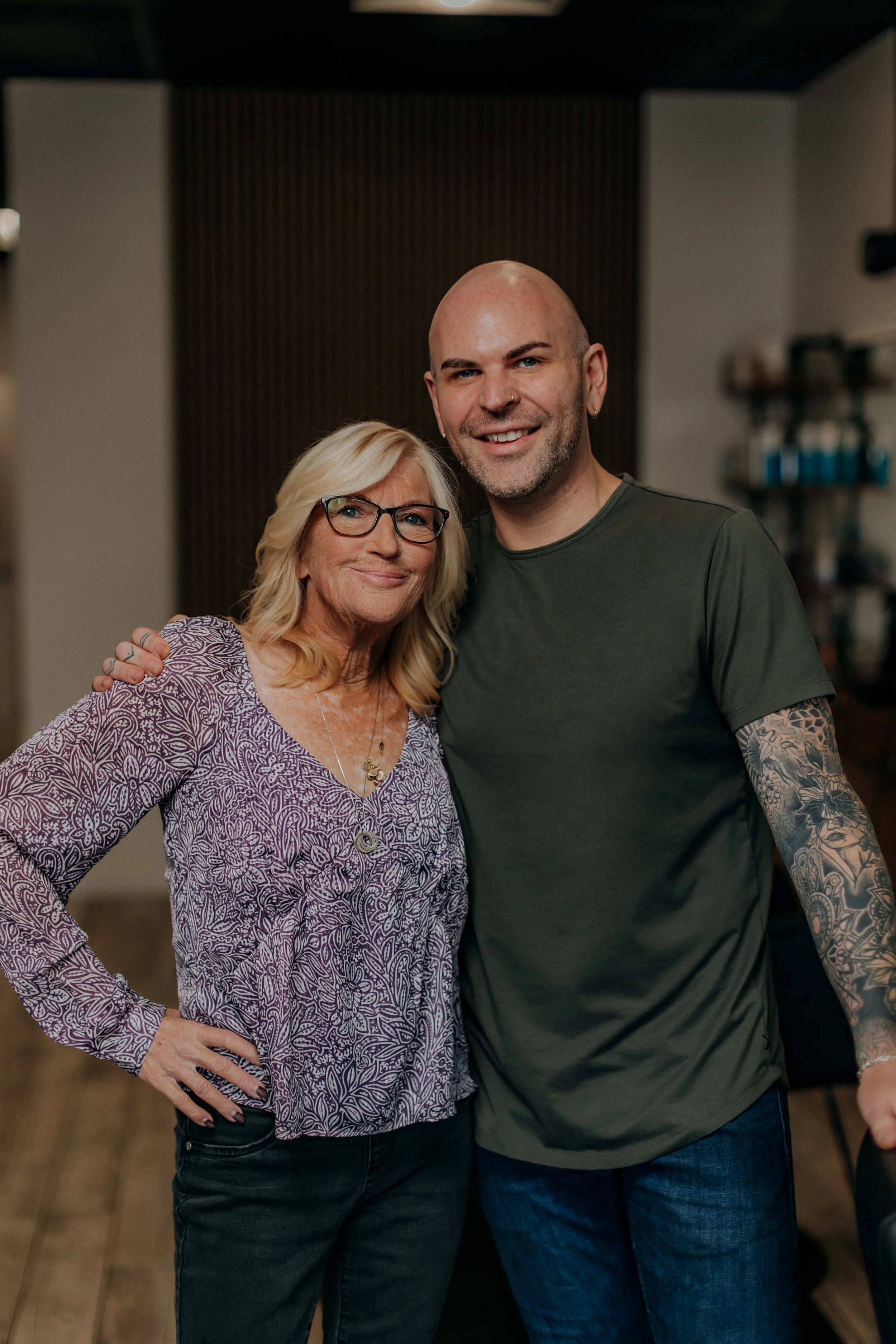 A smiling woman with glasses and blonde hair standing next to a smiling man with tattoos on his arm and a bald head, both posing for the camera indoors.