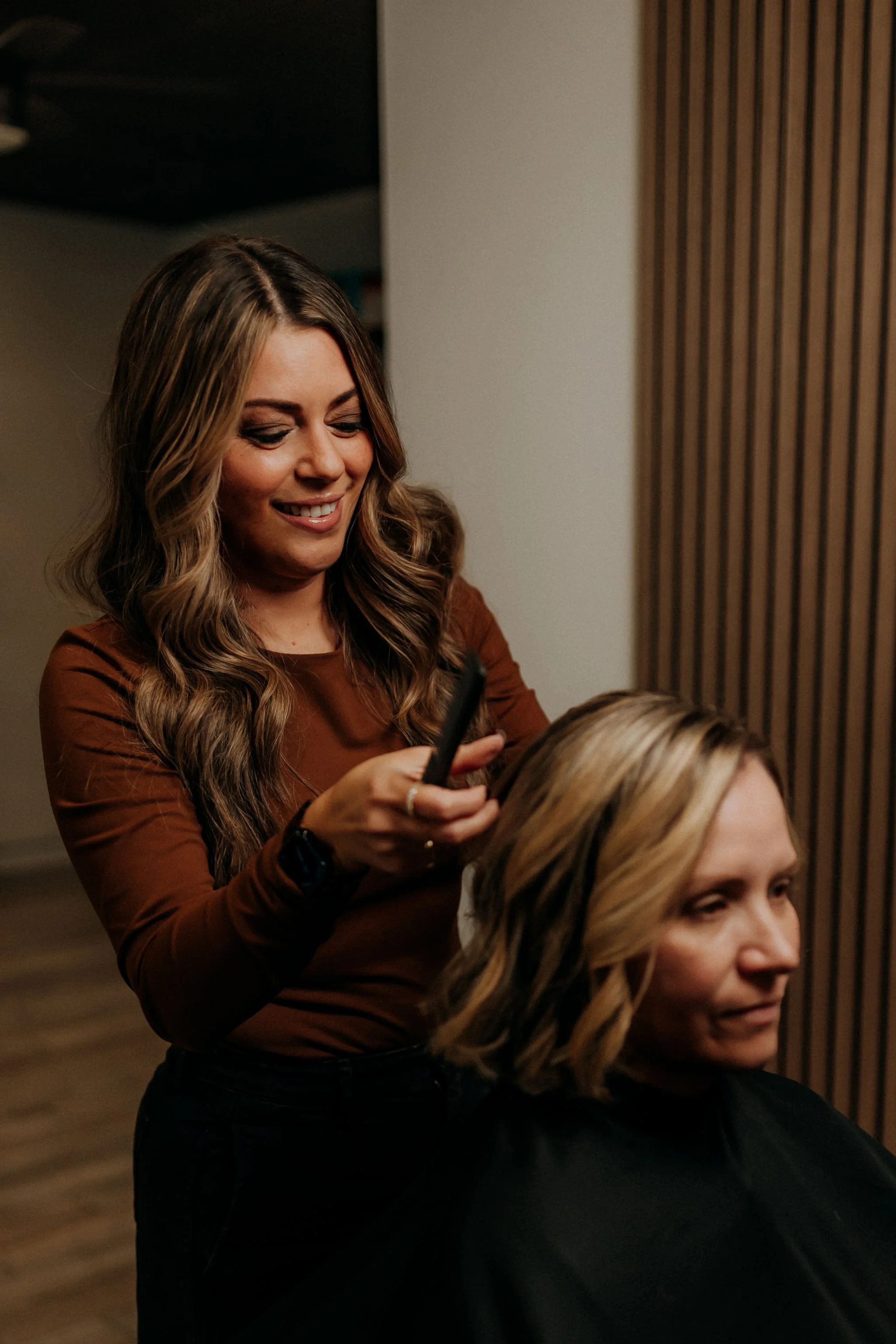 A woman with wavy brown hair smiling while styling another woman’s blonde hair with a curling iron in a salon.