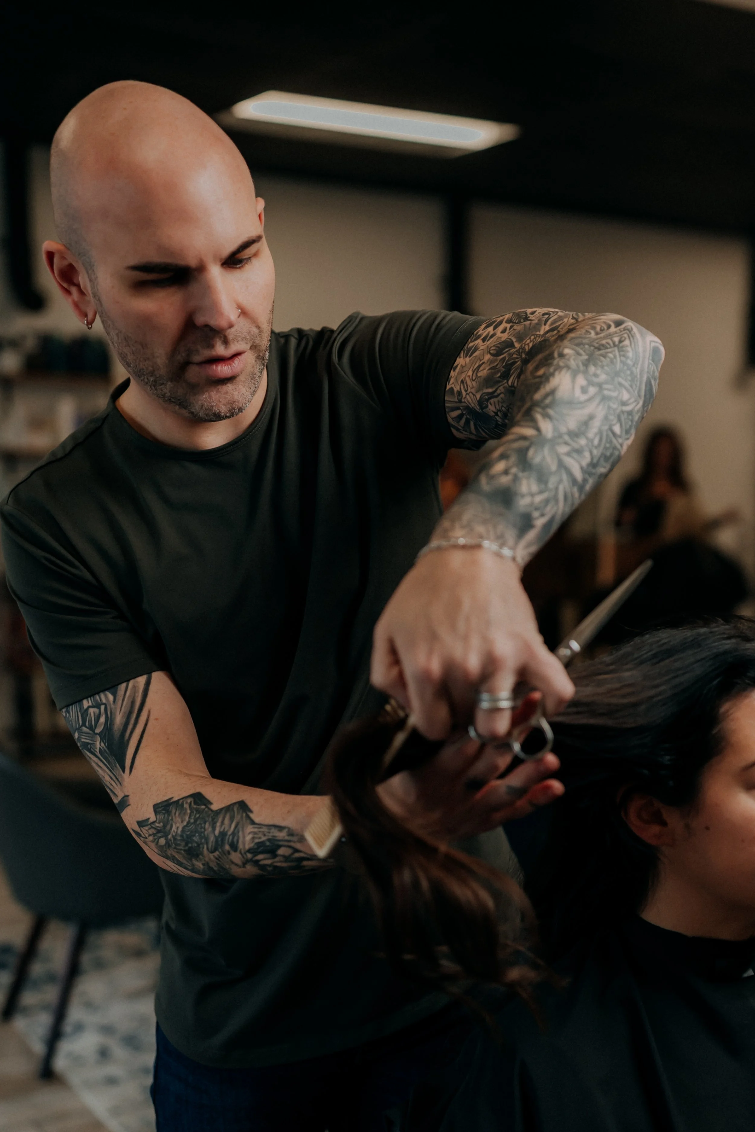 A bald male hairstylist with tattoos on his arms cutting a woman's hair in a salon.