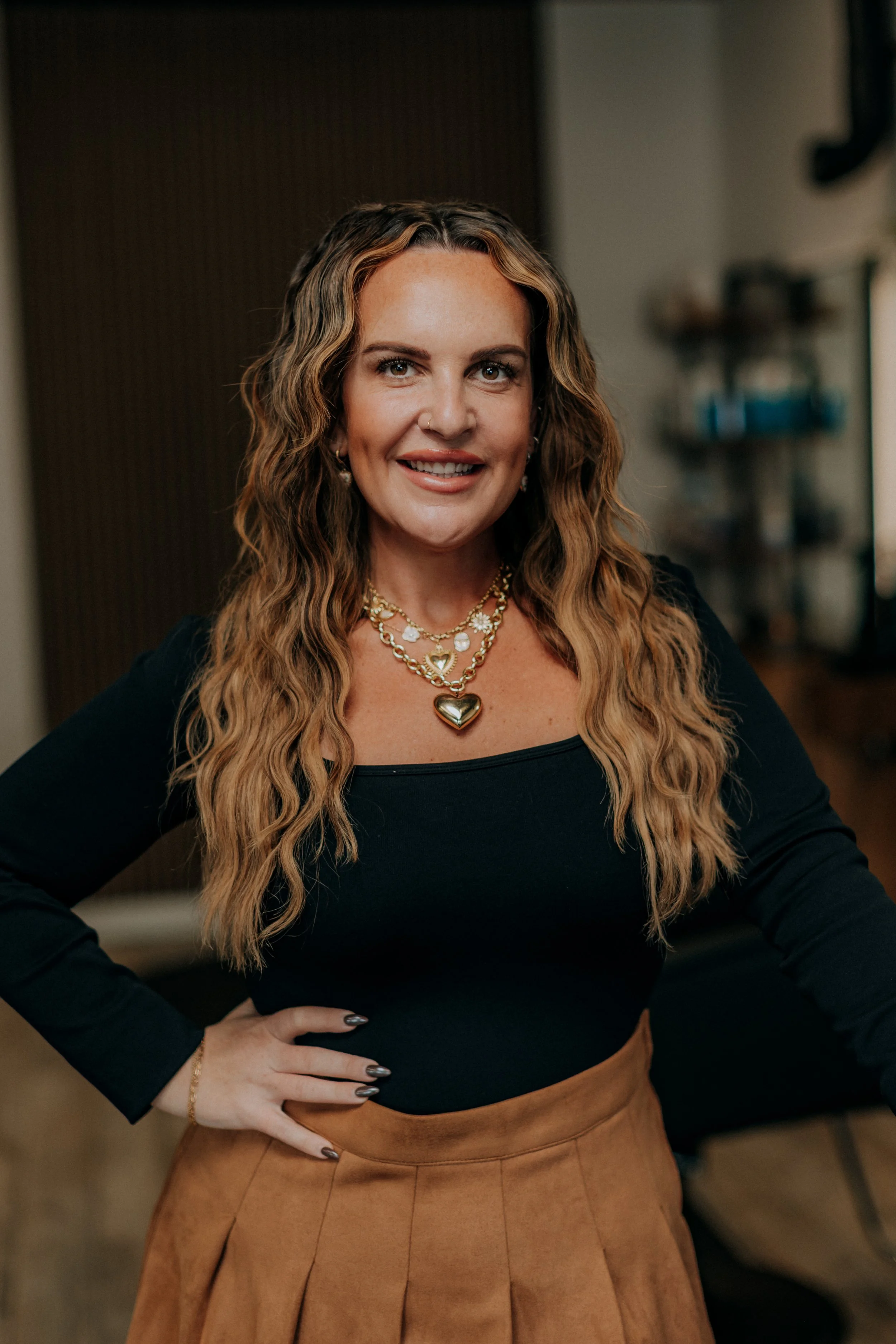 A woman with long, wavy hair, wearing a black top and a tan skirt, smiling and posing with her hand on her hip in an indoor setting.