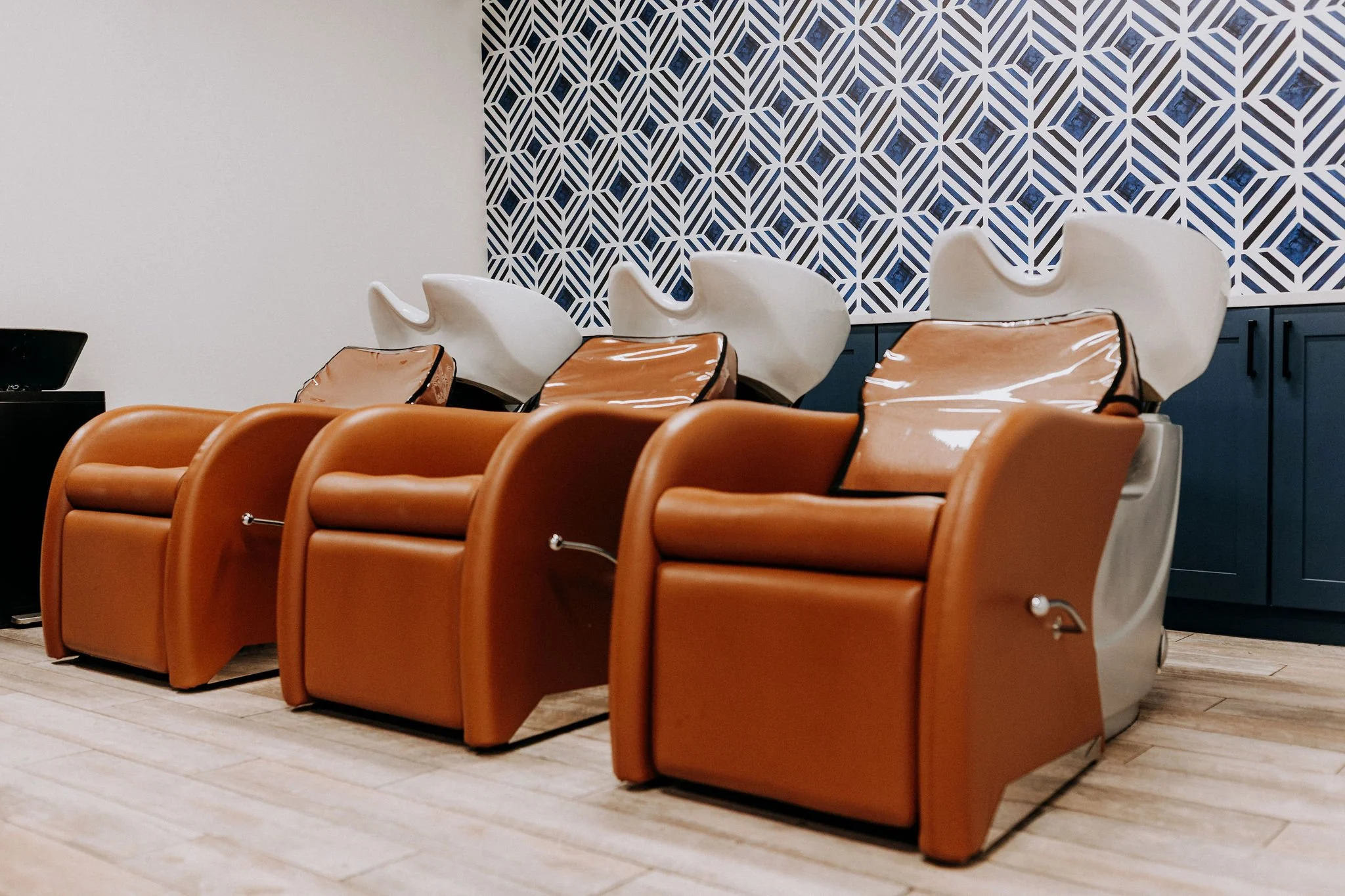 Three brown leather pedicure chairs with white and beige hair steamer units in a salon with a patterned blue and white accent wall and light-colored wooden flooring.
