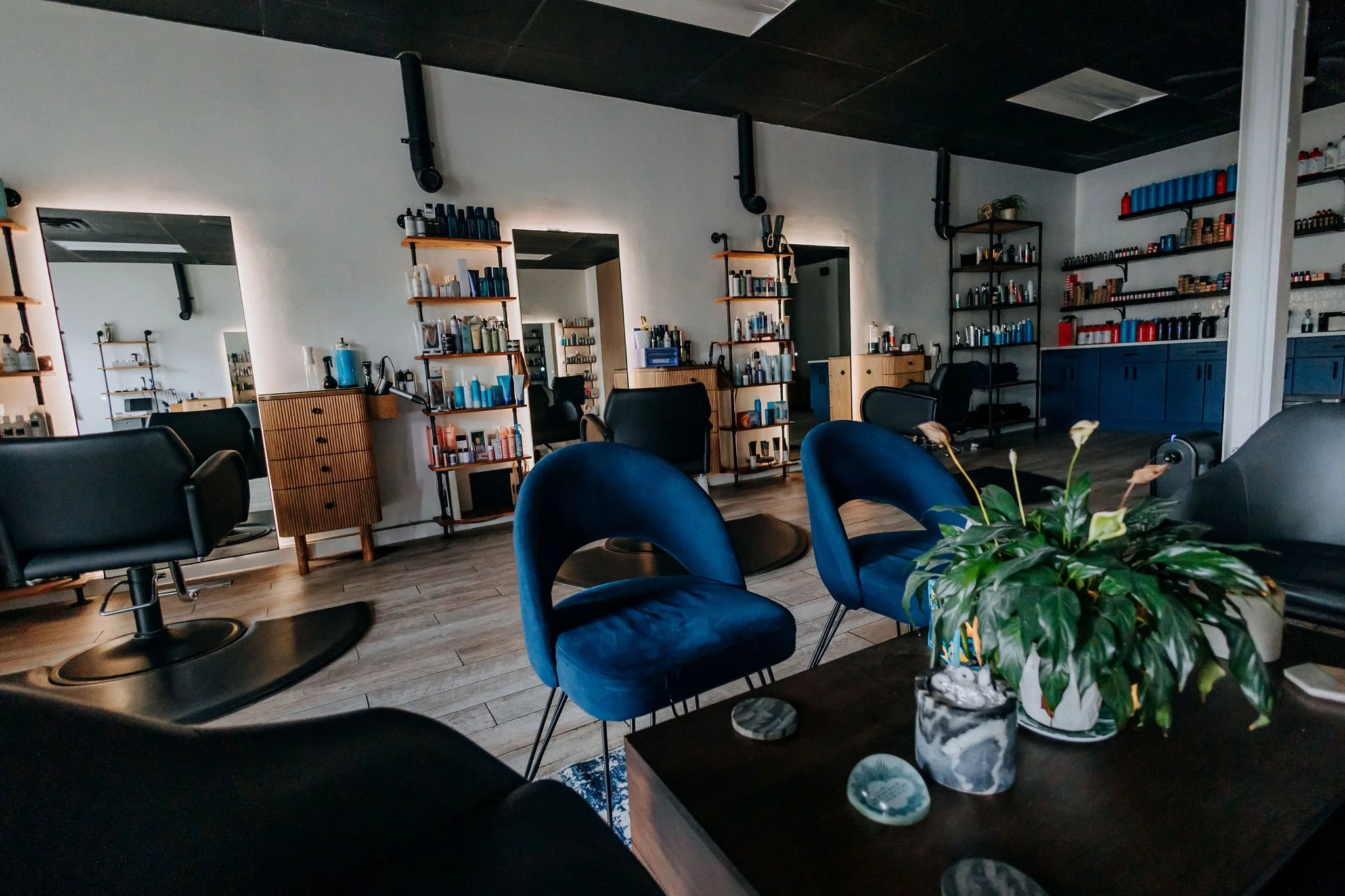 Interior of a modern salon with black chairs, blue velvet armchairs, and wooden shelving with hair and beauty products. There are large mirrors on the wall and a dark ceiling with industrial ductwork.