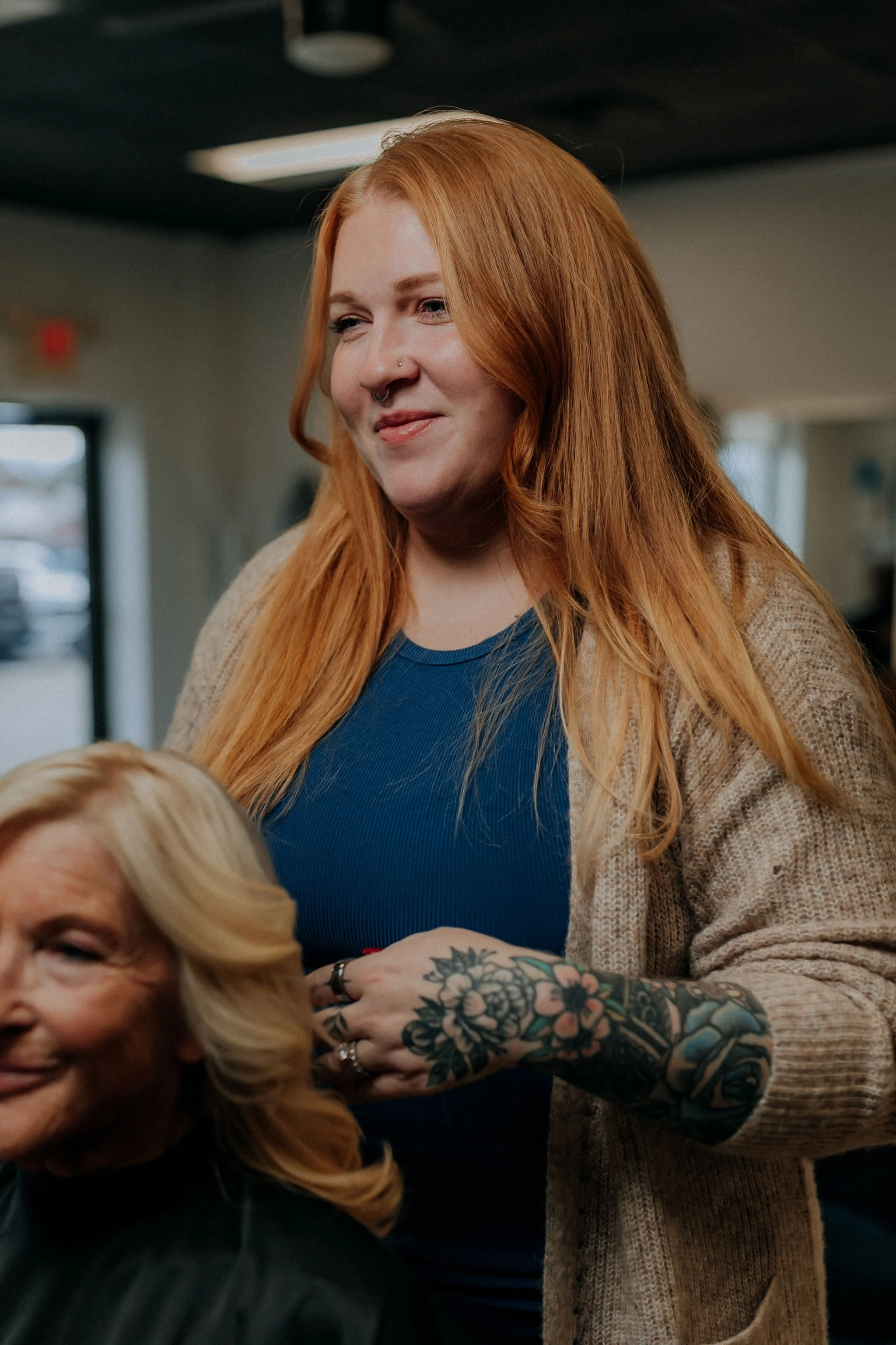 A woman with long red hair and tattoos on her arm smiling, standing next to an older woman with blonde hair, in a salon or similar setting.