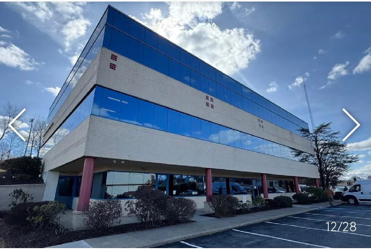 Modern three-story office building with glass windows, pink support columns, and a parking lot in the foreground, under a partly cloudy sky.