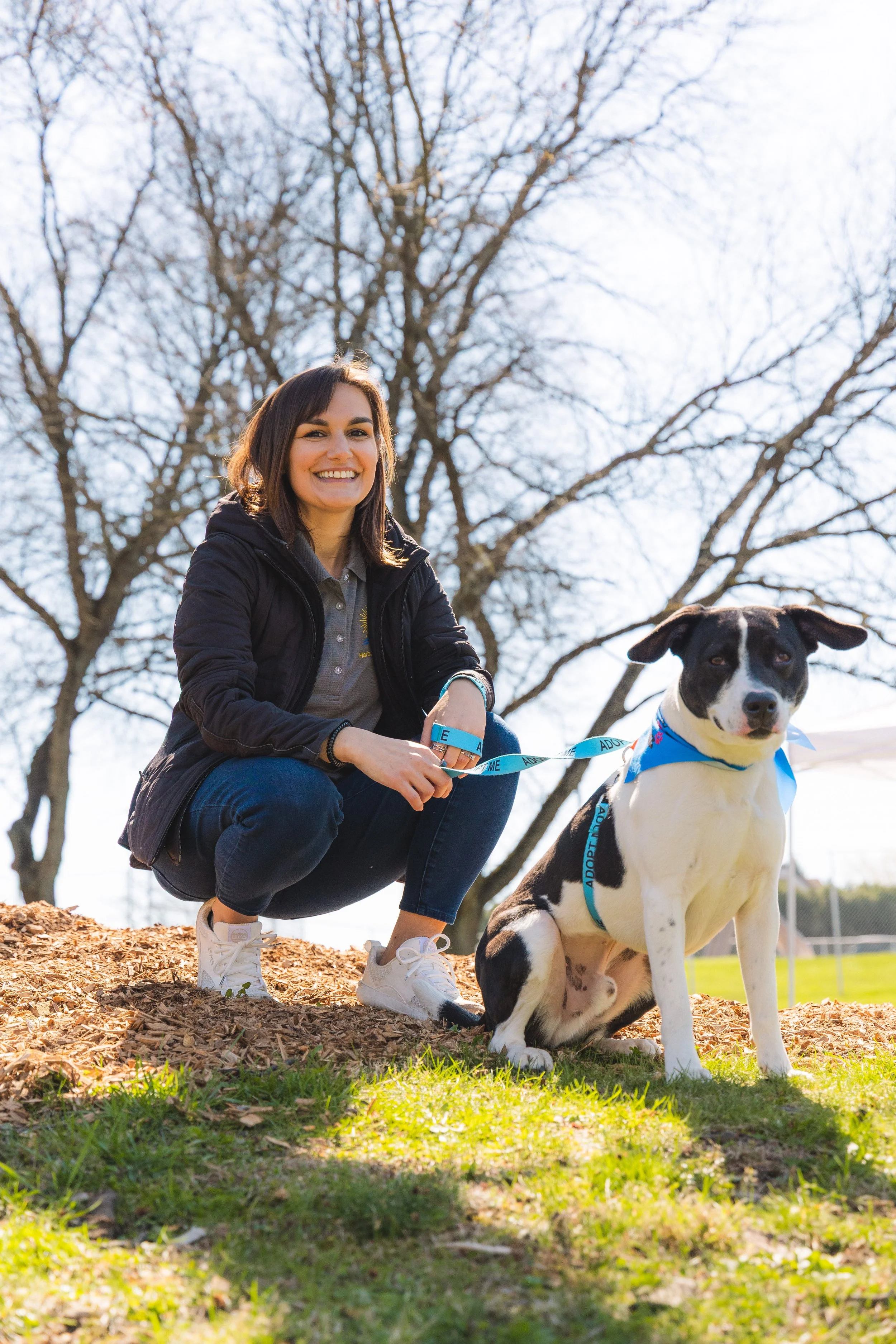 A young woman crouching next to a black and white dog on a leash in a park with trees in the background.