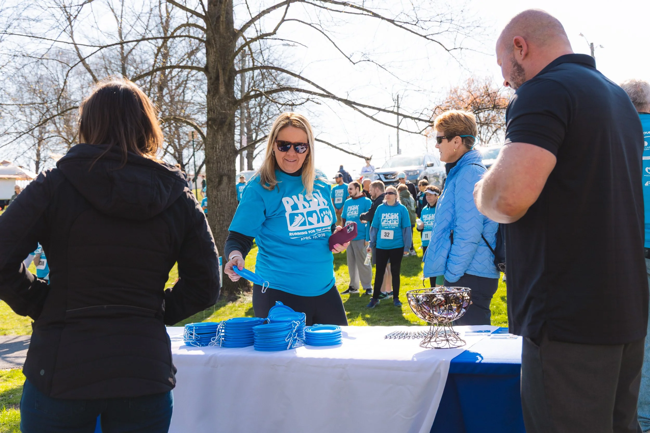 People gathering outdoors at a charity or awareness event, wearing blue shirts, near a table with blue wristbands and a wire basket of medals or keychains, under a large tree with bare branches.
