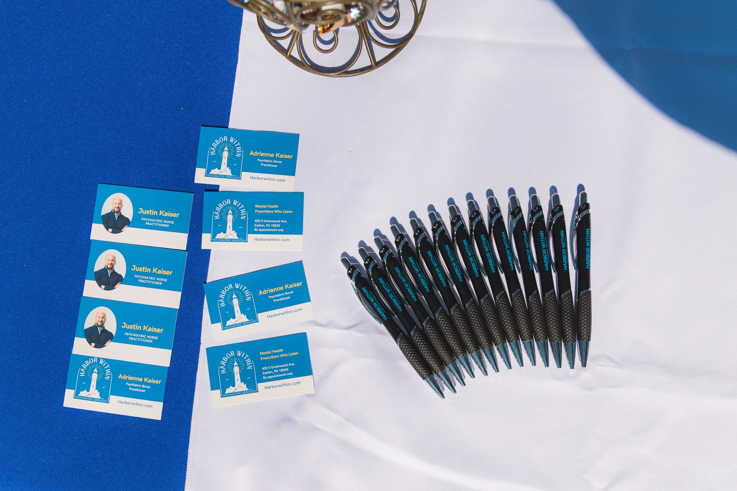 Business cards and pens arranged on a table with a blue and white tablecloth, featuring name tags for Justin Kaiser and Adrienne Kaiser from Harbor Within, a psychiatric services provider.
