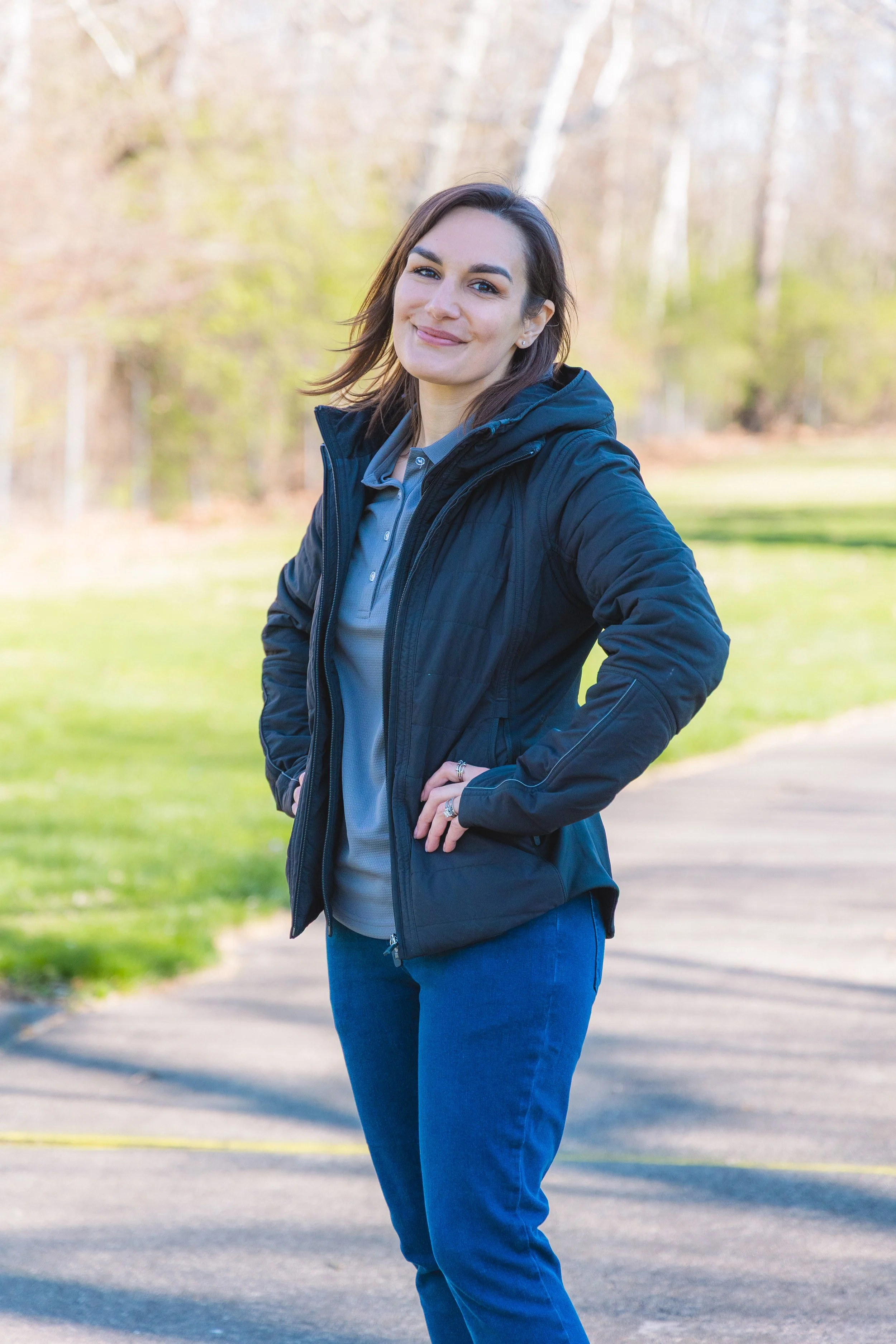A woman with shoulder-length brown hair, wearing a navy jacket and blue jeans, standing outdoors in a park with a background of trees and grass, smiling at the camera.