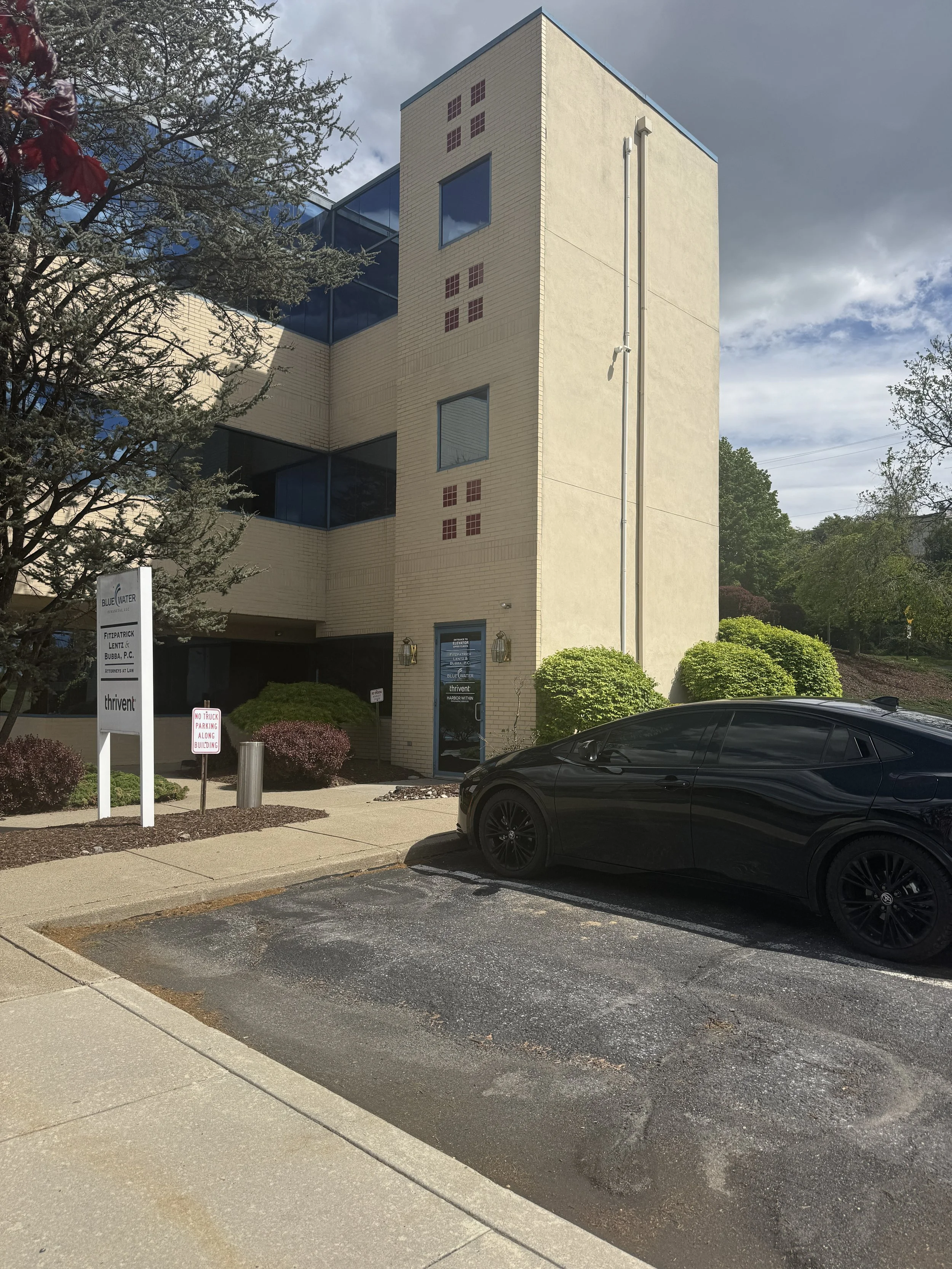 A modern office building with beige brick walls, large glass windows, and red decorative elements. There is a black car parked in front, and trees and shrubs are visible around the building. The sky is partly cloudy with dark clouds.
