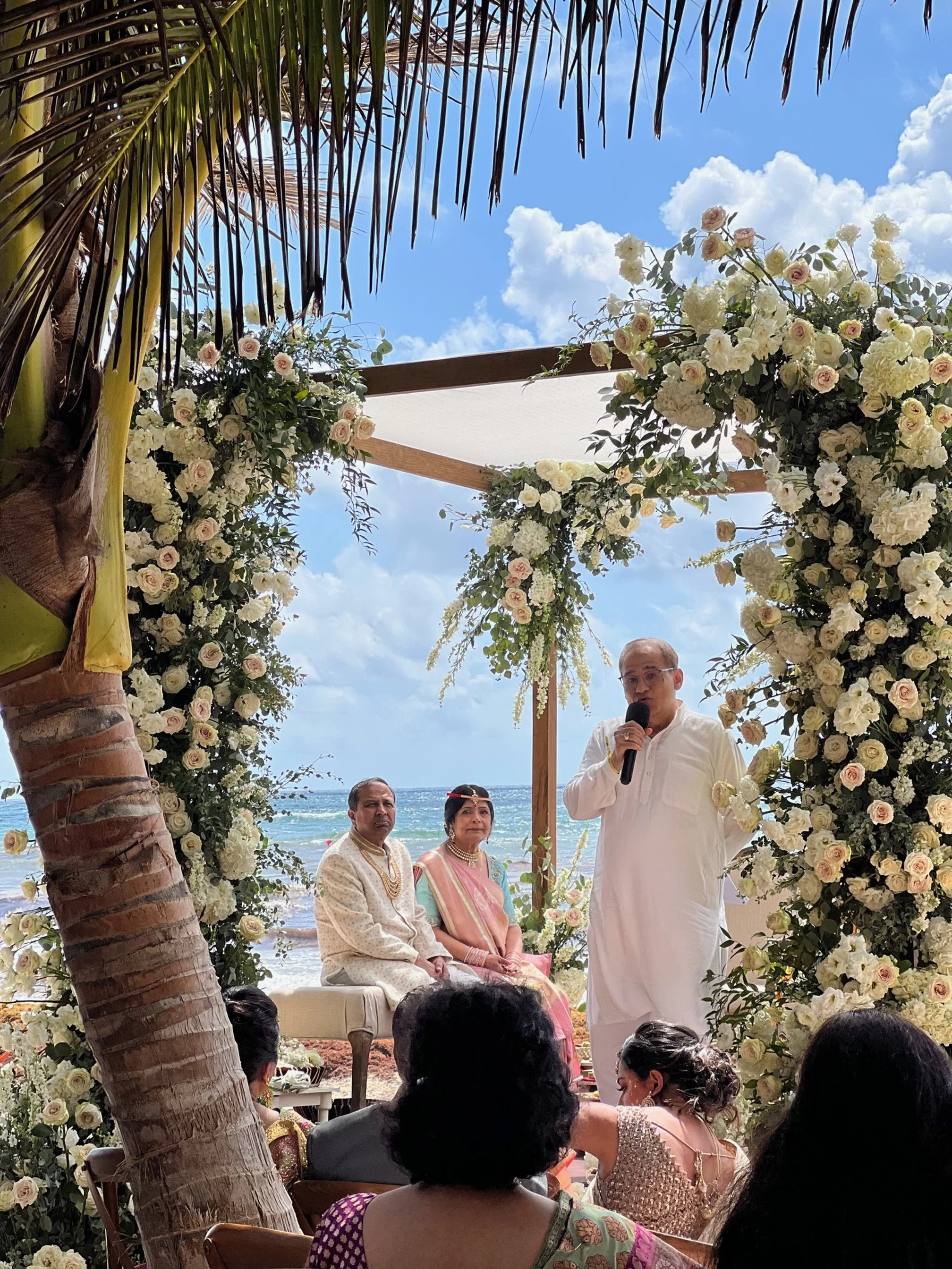 A wedding ceremony on a beach with a floral arch, a speaker holding a microphone, and the couple seated on a sofa, with the ocean and blue sky in the background.