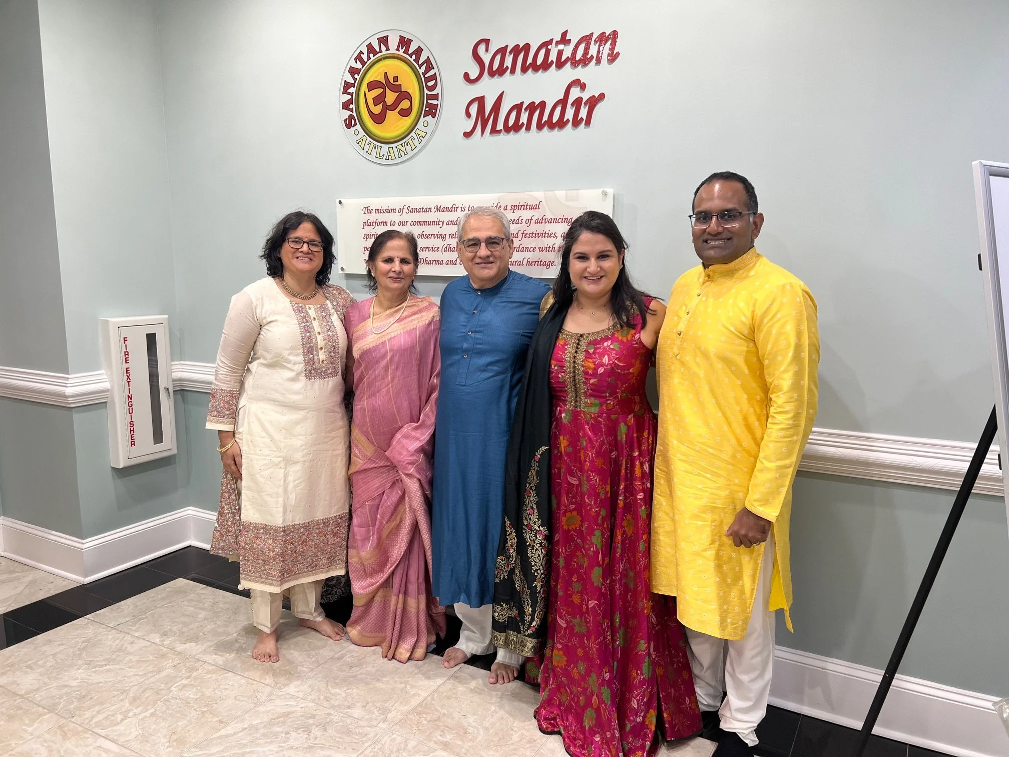 Five people standing together in front of a sign that says "Sanatan Mandir" with a religious symbol at a Hindu temple. They are all dressed in traditional Indian clothing and smiling.