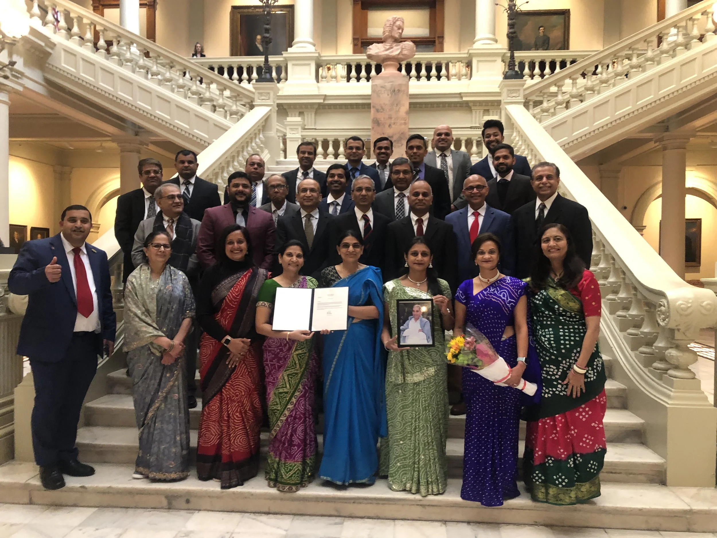 Group of people posing on a grand staircase in an elegant building, some holding framed photos and documents, dressed in formal and traditional attire.