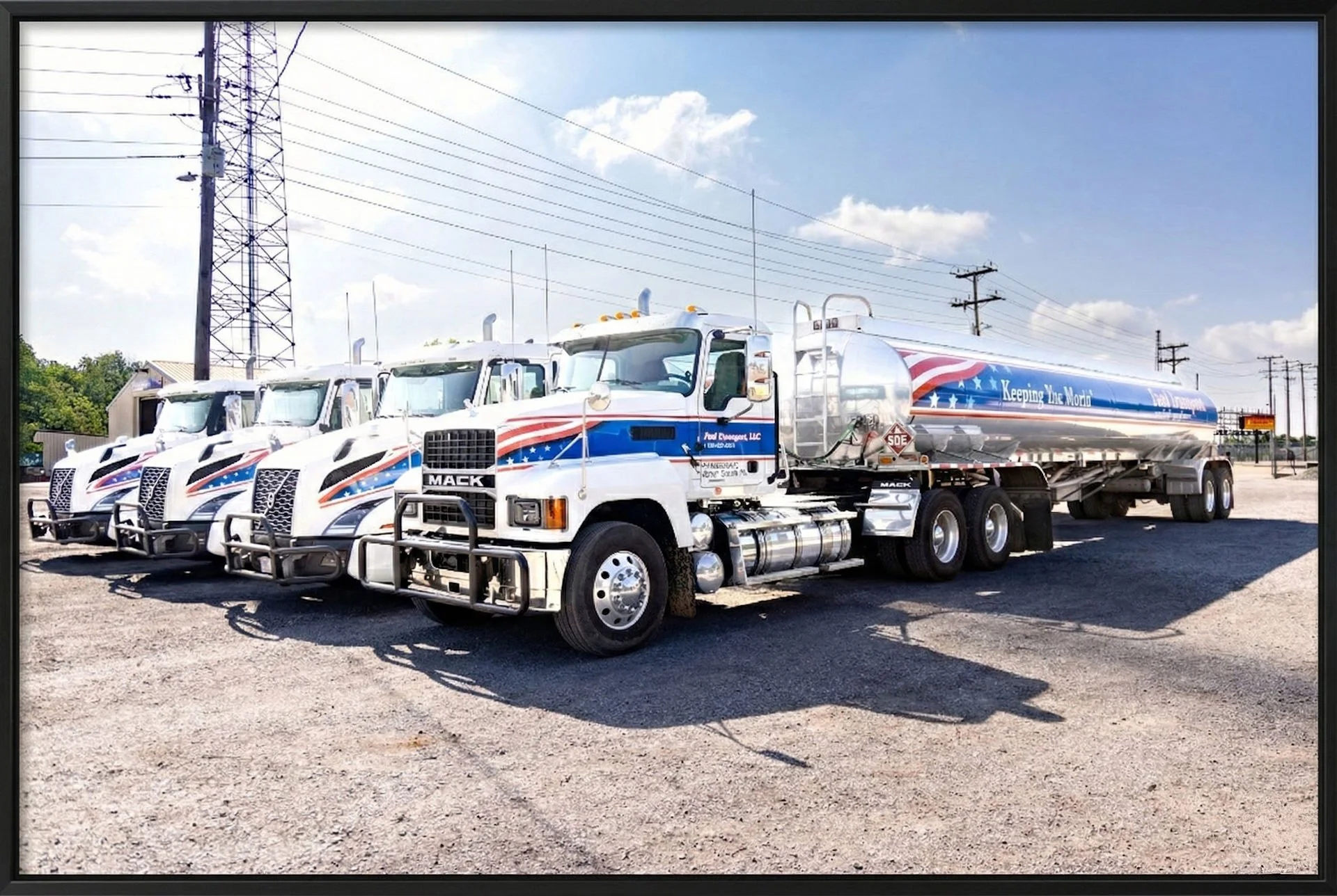 Lineup of four white tanker trucks with patriotic red, white, and blue decals, parked on gravel lot with power lines and small buildings in the background.
