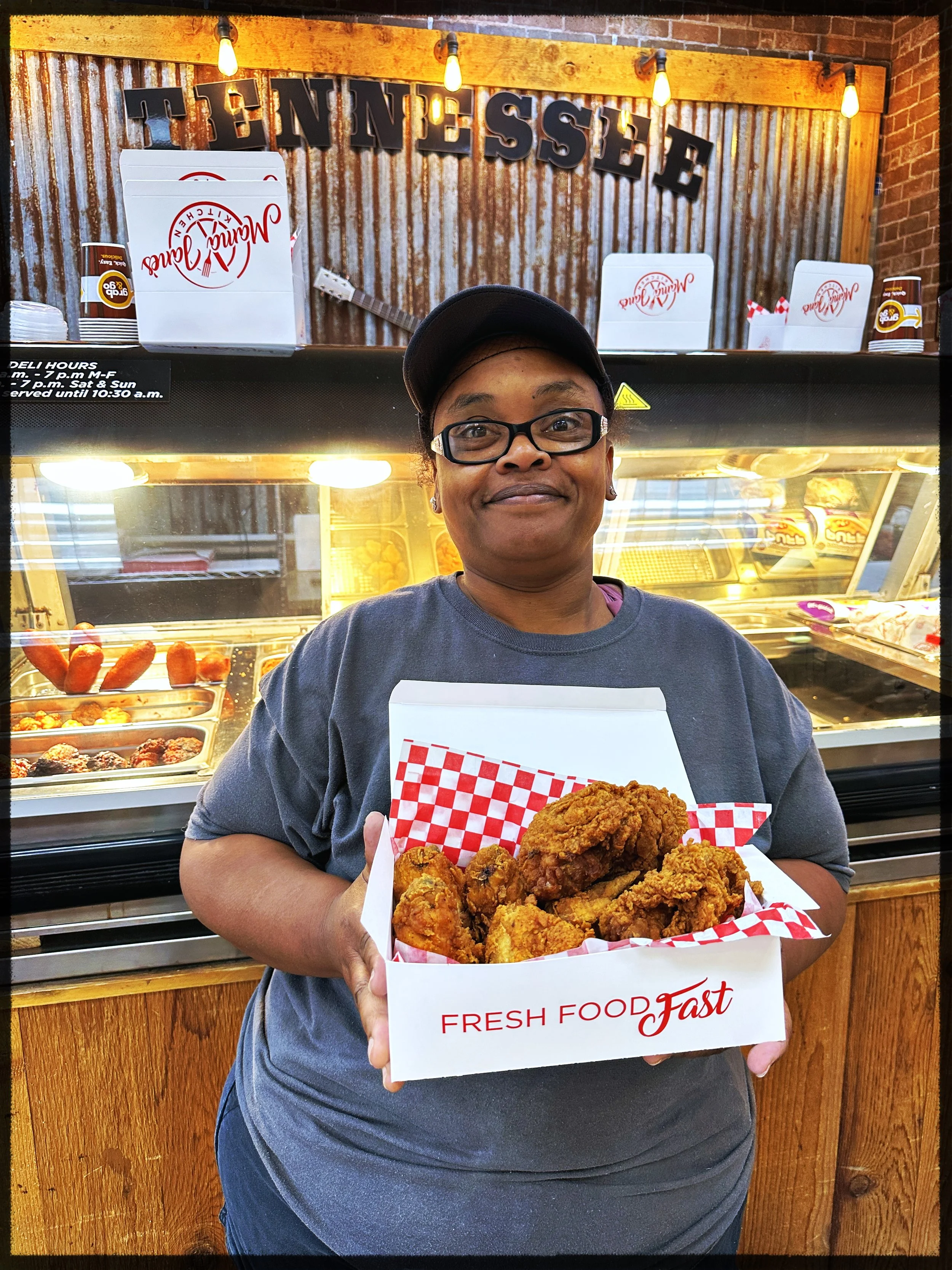 A woman wearing glasses and a black cap holding a white box with fried chicken pieces inside. She stands in front of a food display case at Tennessee restaurant. Decor includes a rustic sign and lights.