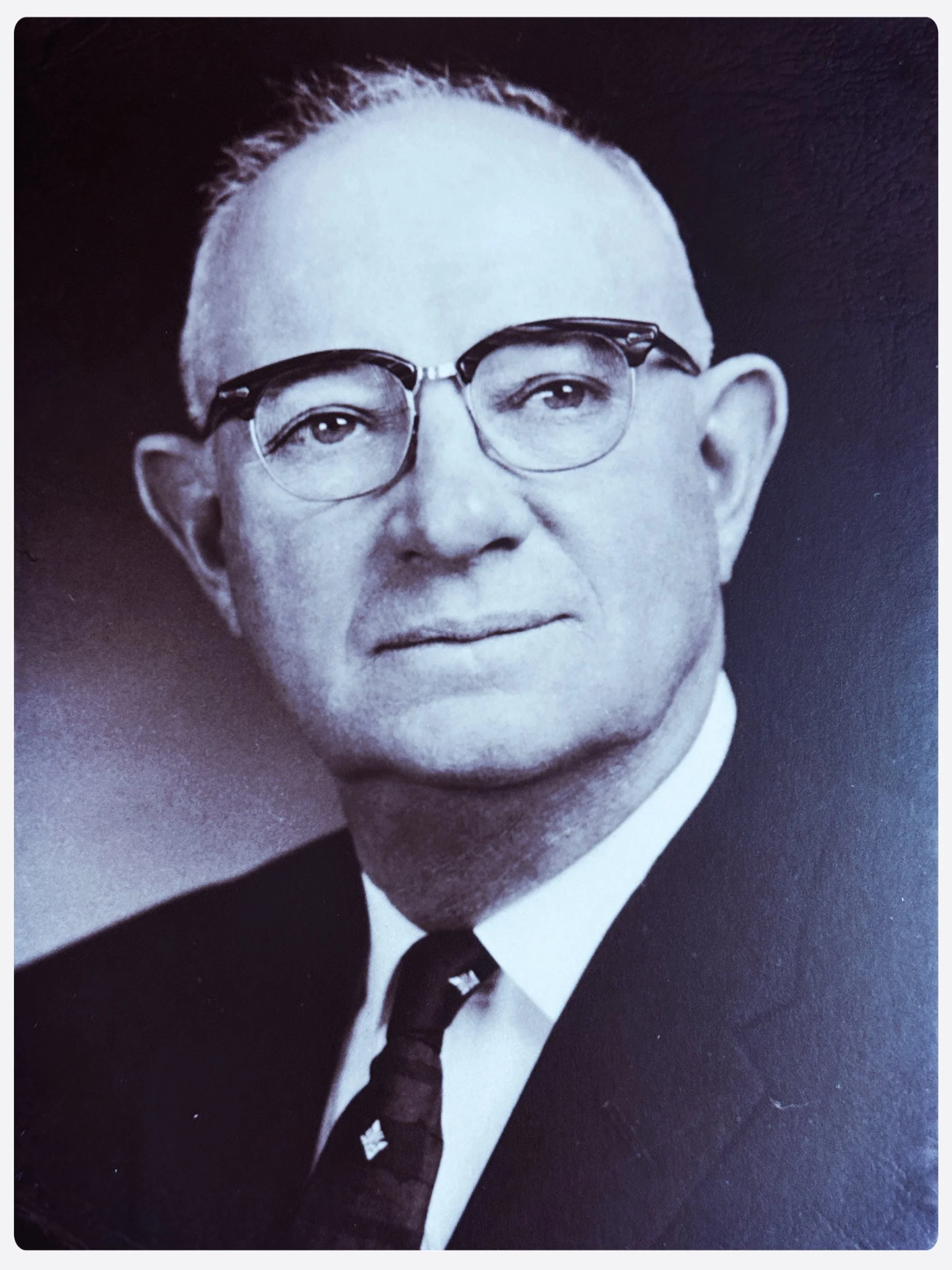 Black-and-white portrait of our founder  E.M. Adcox, wearing glasses, a suit, and a patterned tie.