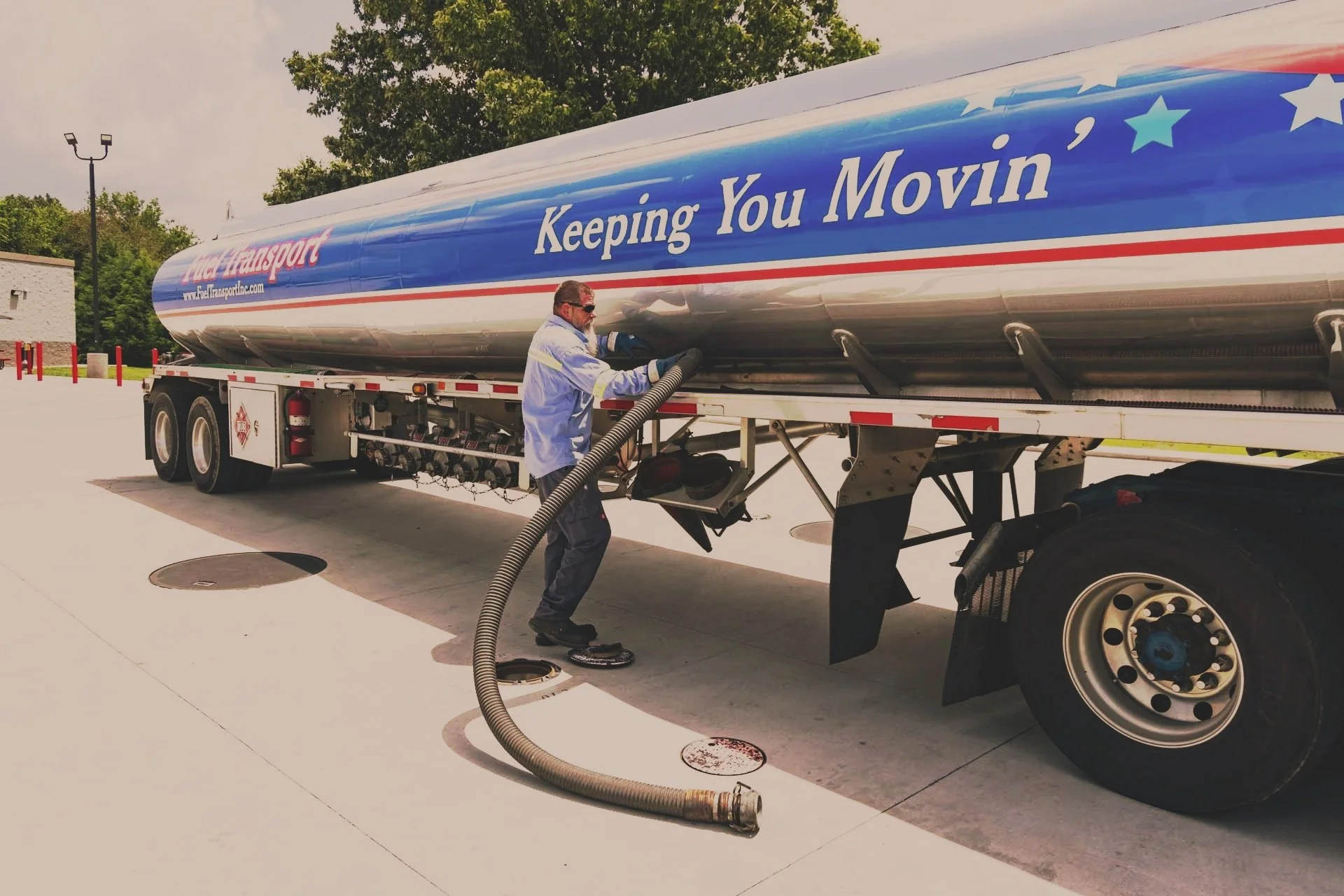 A man in work clothes vacuuming the underside of a large fuel transport truck with a blue, red, and white design that reads, "Keeping You Movin'."