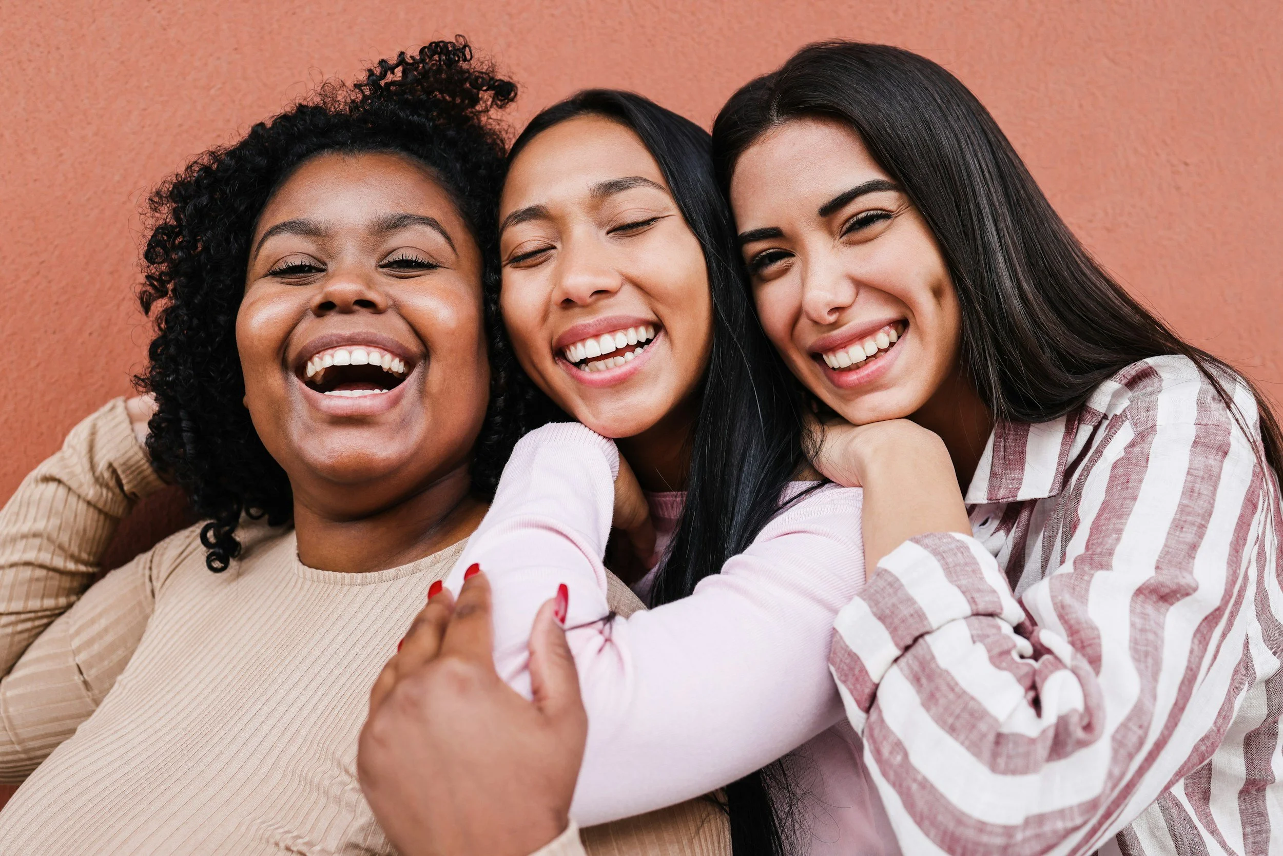 Three women joyful and smiling, hugging each other closely against a coral background.