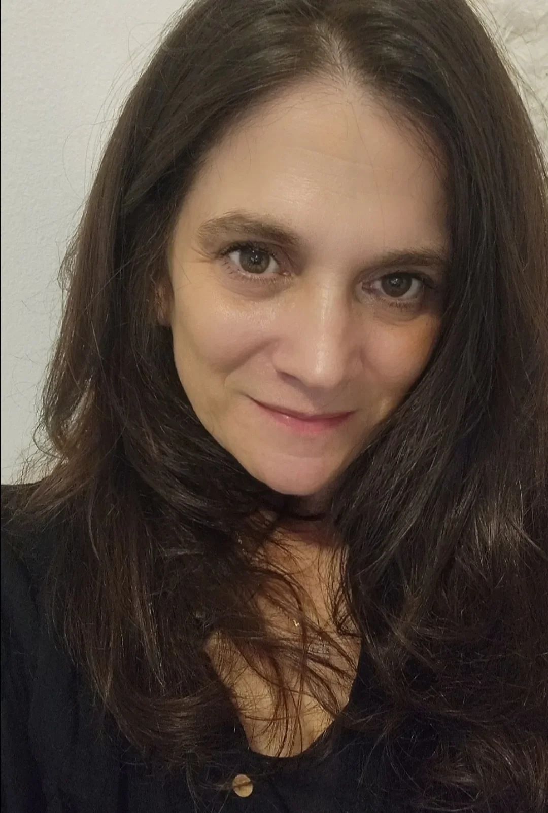 Close-up of a woman with long, dark brown hair and light-colored eyes, smiling softly at the camera. She is wearing a black top and is positioned against a plain light background.