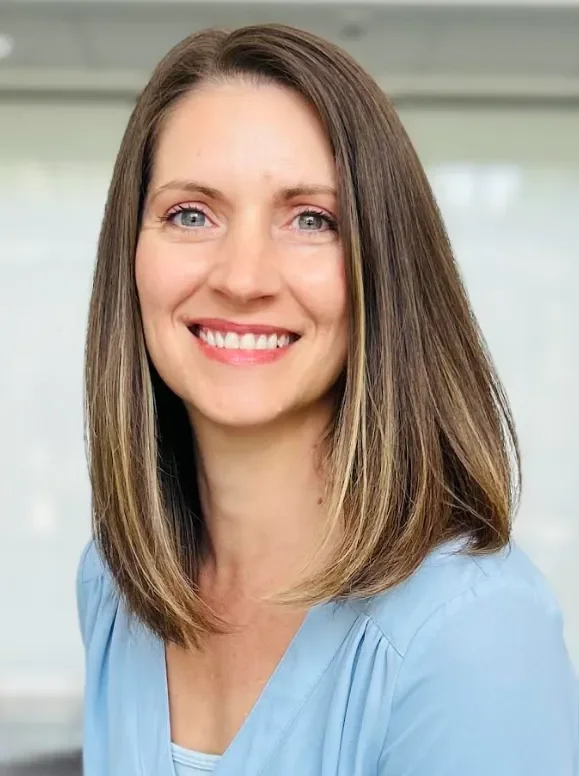 Close-up of a woman with shoulder-length light brown hair, blue eyes, and a warm smile, wearing a light blue top, in a bright indoor setting.