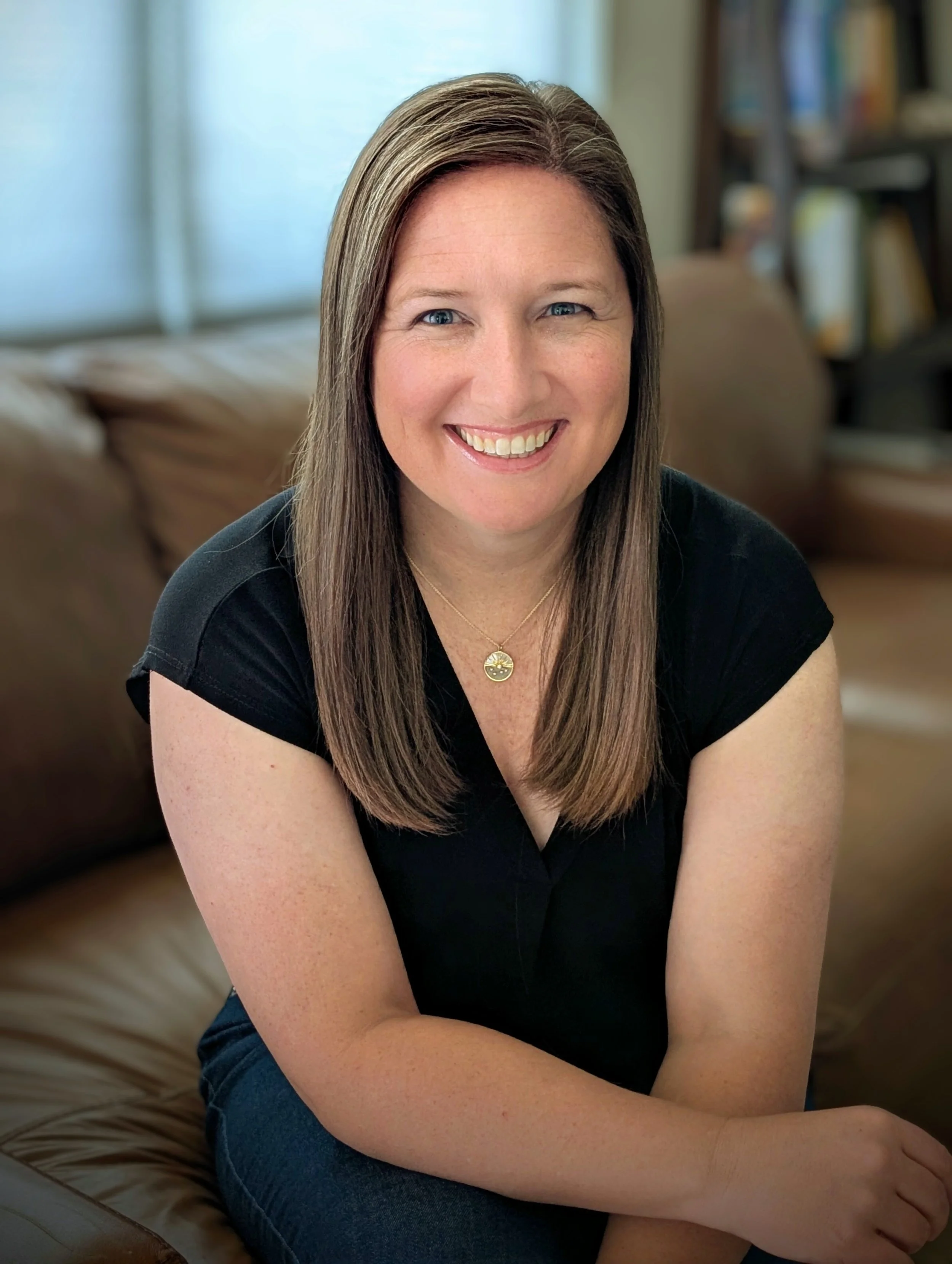 A woman with long brown hair and blue eyes sitting on a brown leather couch in a room with bookshelves in the background, smiling at the camera.