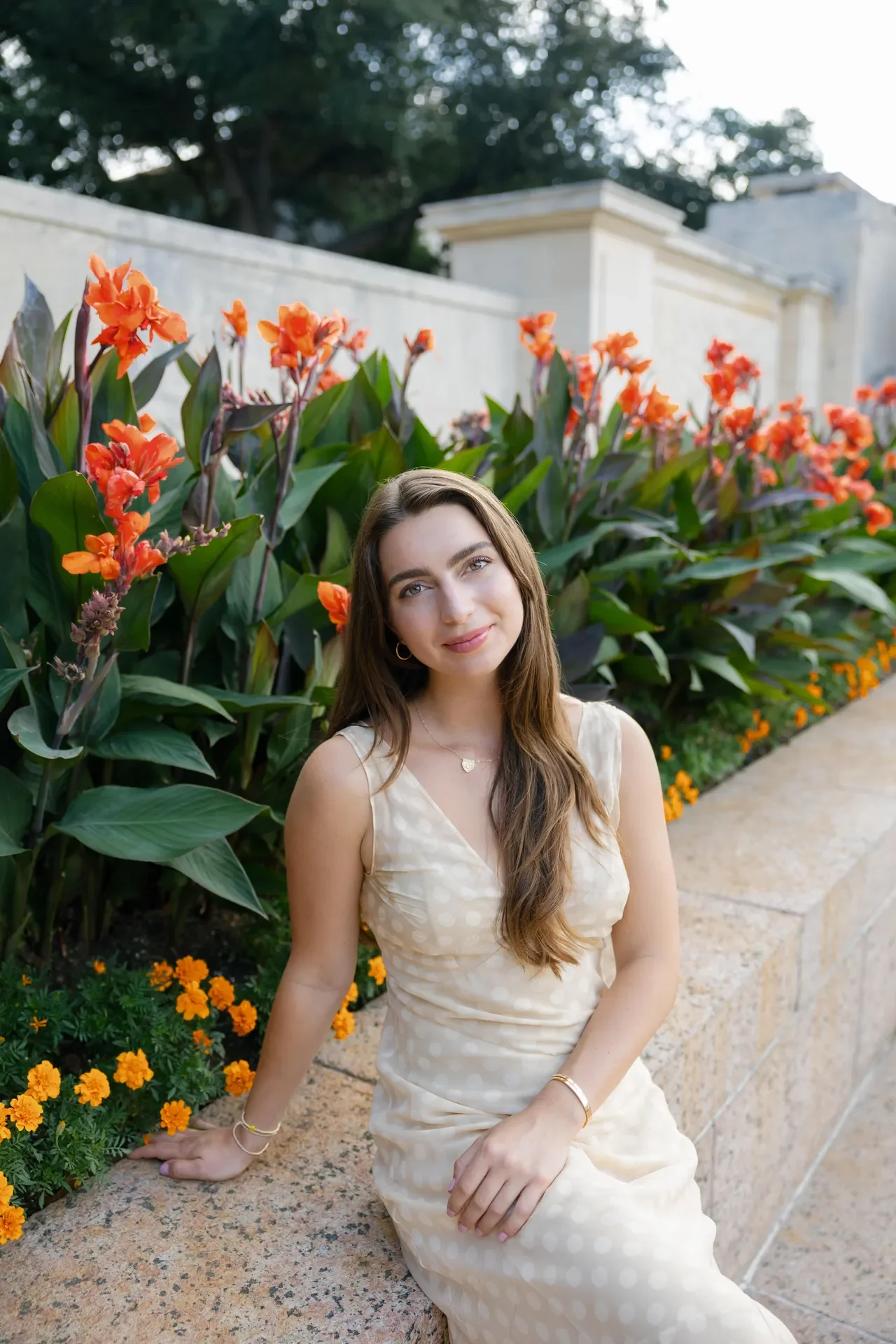 A young woman with long brown hair and light skin sitting on a stone ledge in front of a flower garden, smiling at the camera. She is wearing a cream-colored dress with a polka dot pattern, gold jewelry, and has a relaxed posture.