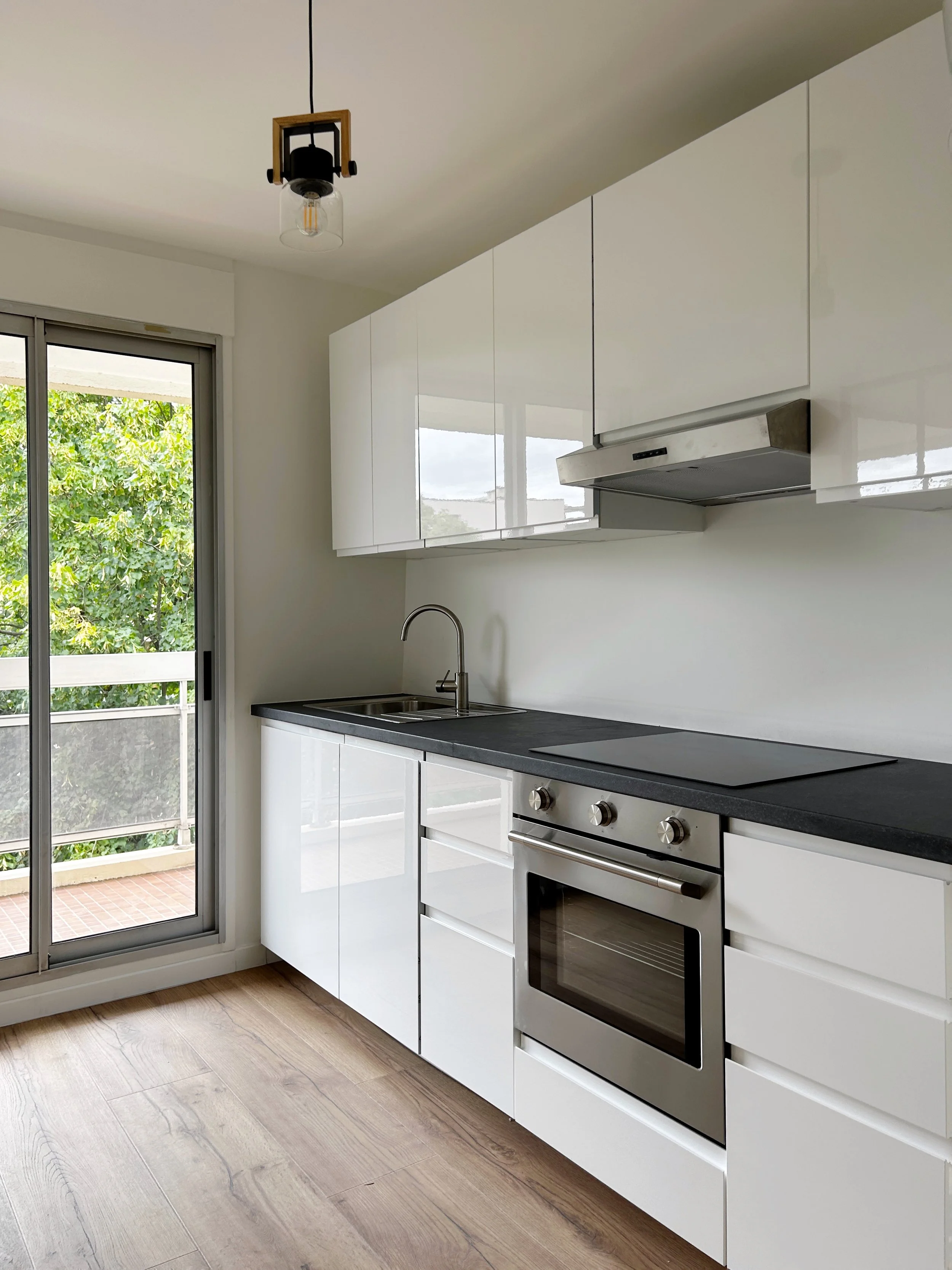 Cuisine moderne avec four électrique, hotte aspirante, évier, et porte-fenêtre donnant sur un balcon avec vue sur la verdure.