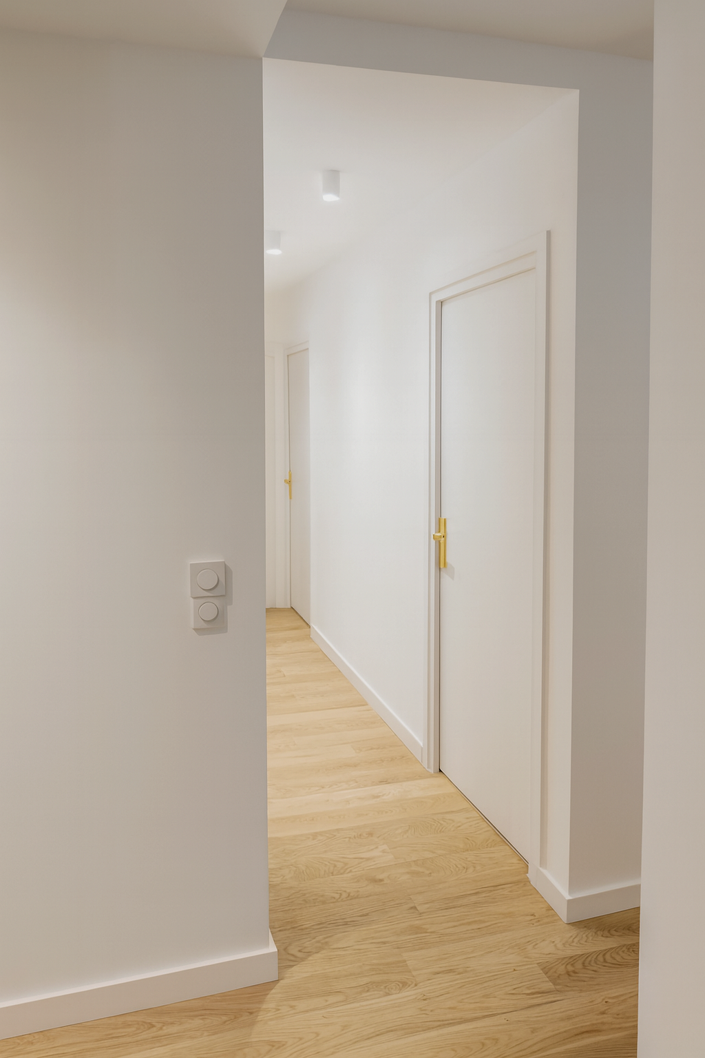 Un couloir dans une maison avec des murs blancs, deux portes blanches avec poignées dorées, et un sol en bois clair.