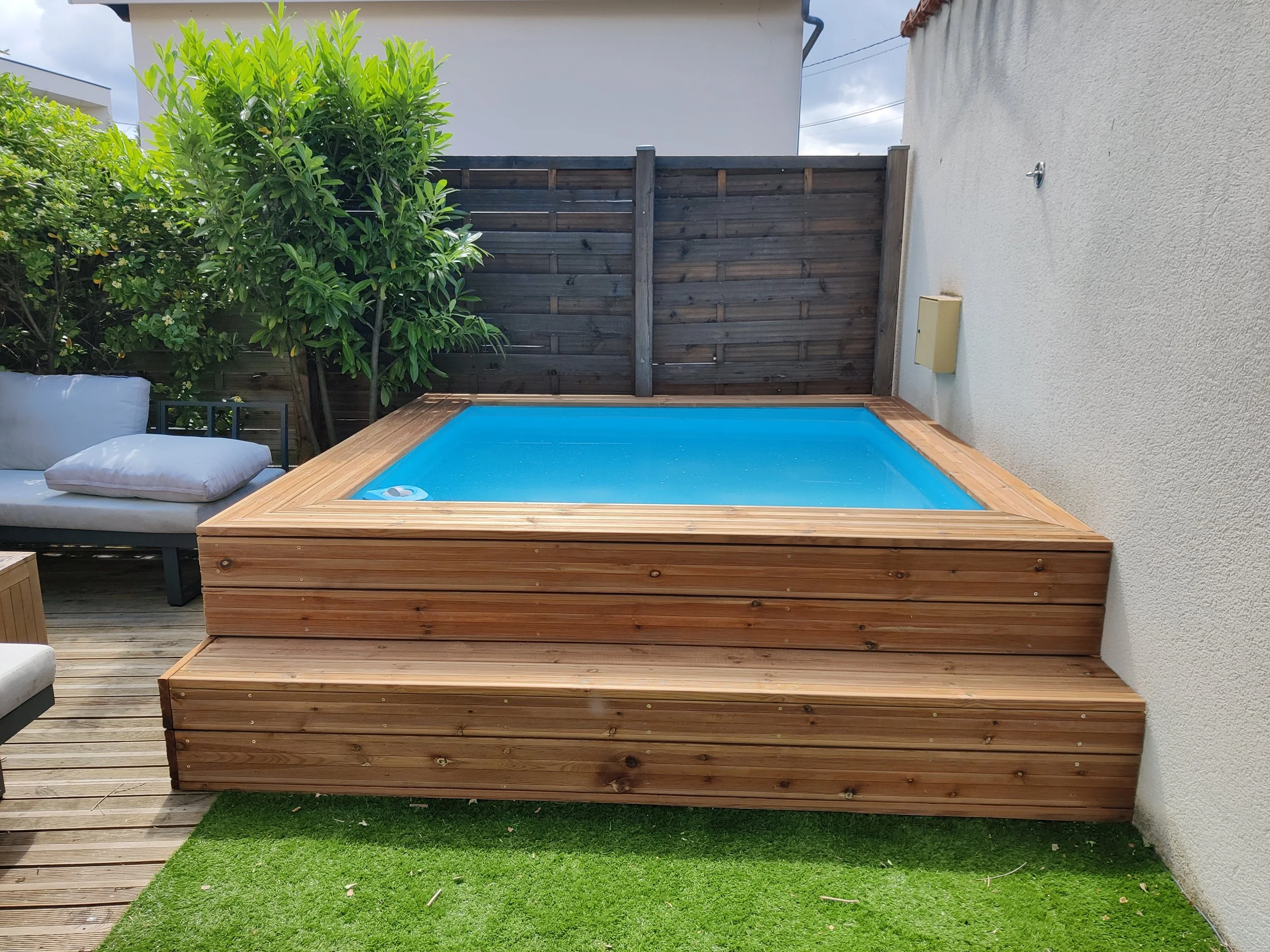 Piscine en bois dans un jardin, avec canapé en extérieur et végétation luxuriante en arrière-plan.