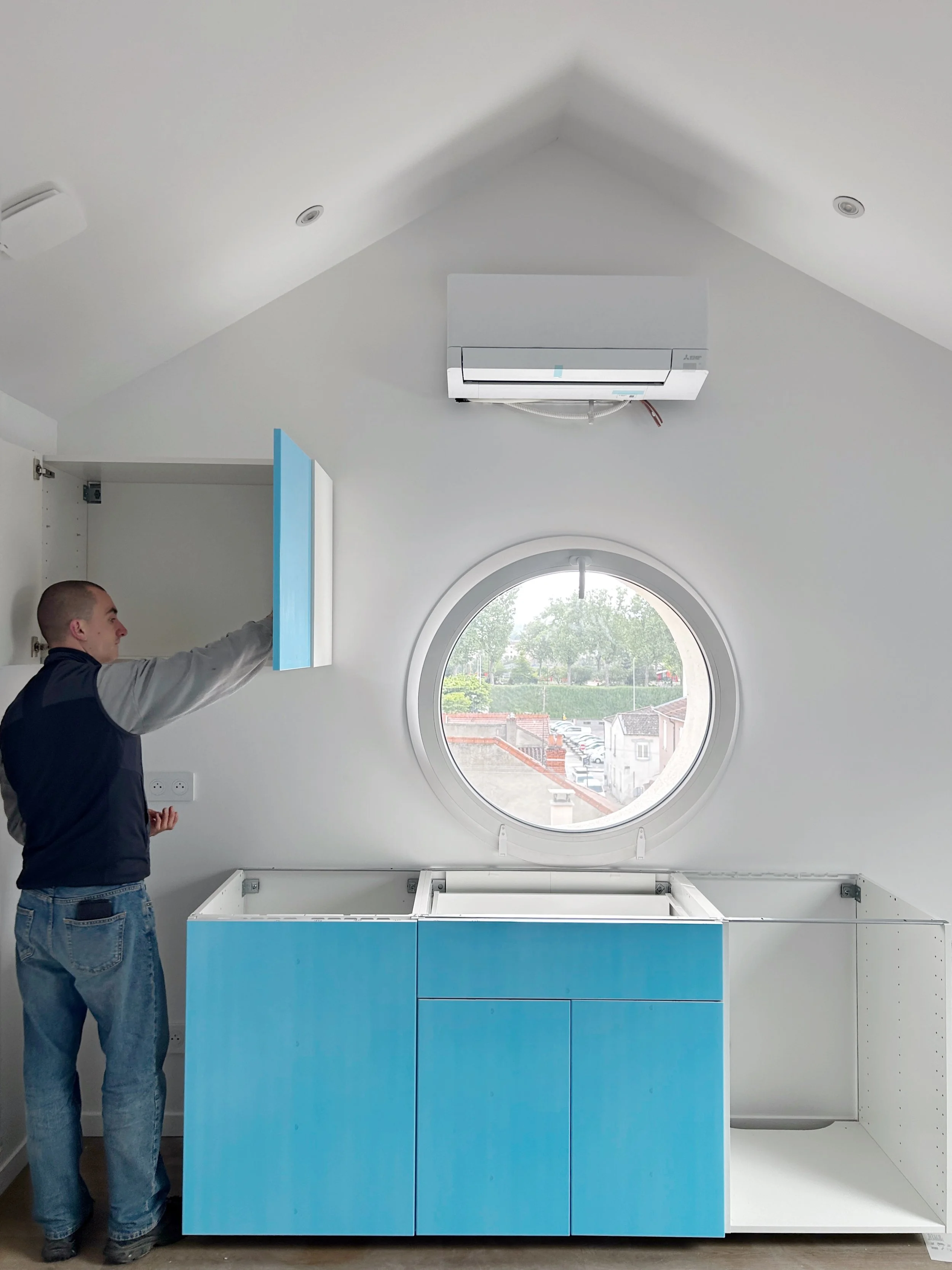 Une personne installe ou inspecte un meuble de cuisine bleu clair avec des tiroirs dans une pièce avec un plafond blanc. Il y a une fenêtre ronde derrière le meuble, et un miroir ou un panneau blanc sur le côté droit.