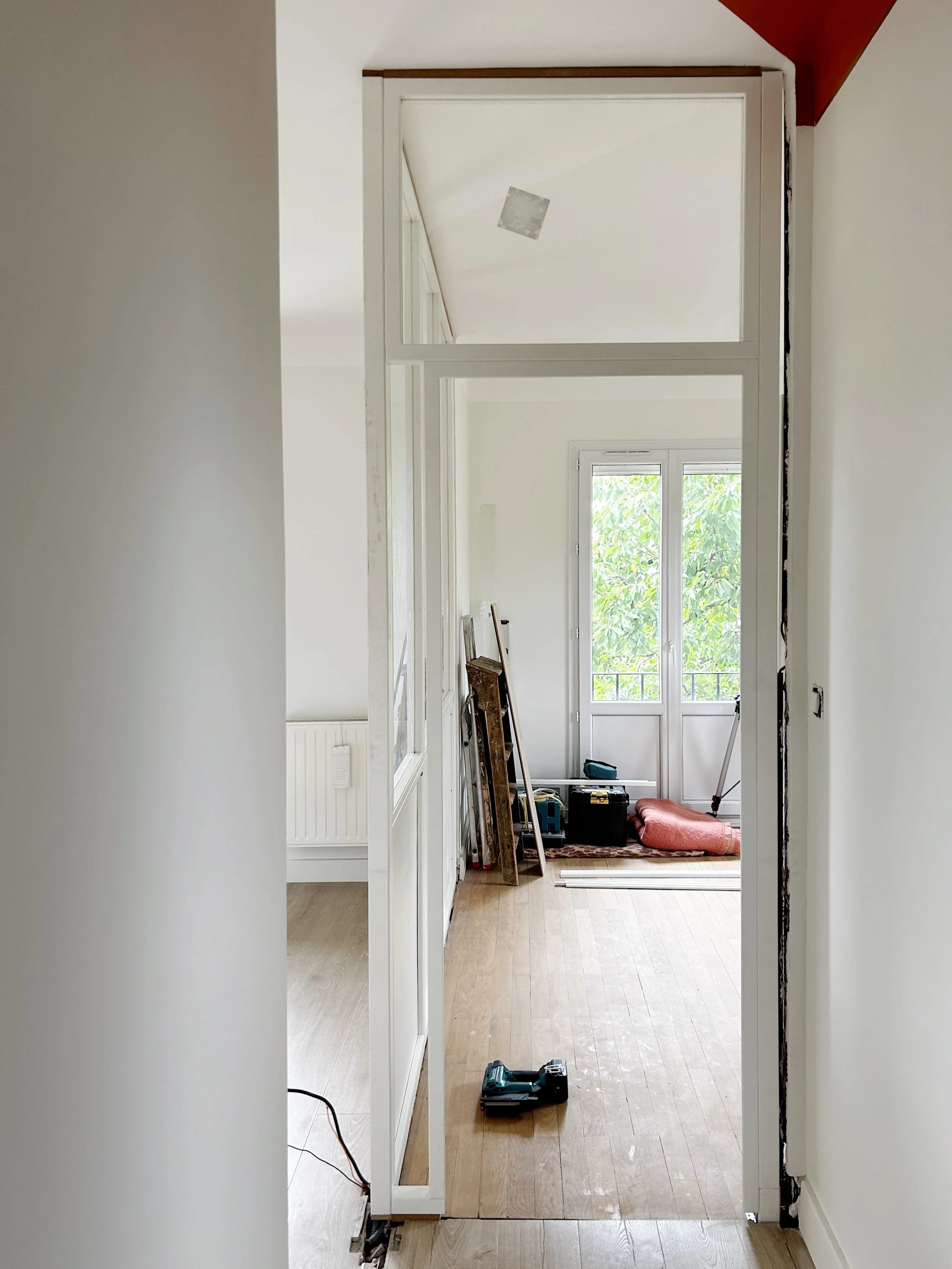 Un couloir en cours de rénovation avec des outils, un escabeau et un matelas dans une pièce lumineuse avec des fenêtres sur le fond.