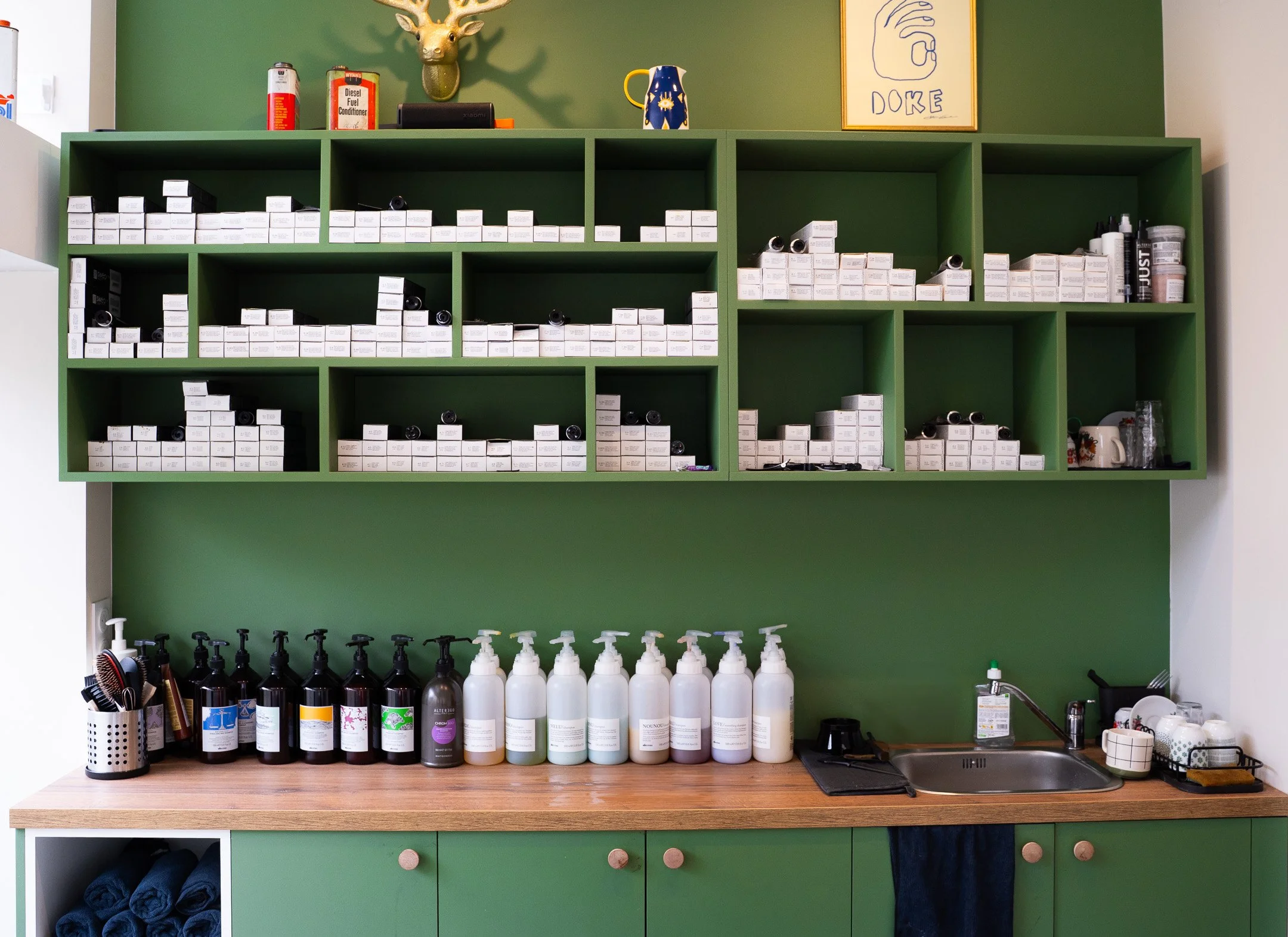 Étagère verte avec divers médicaments et produits de soins, bouteille de savon liquide, évier avec une serviette noire, et plusieurs contenants de savon ou lotion sur le comptoir en bois.