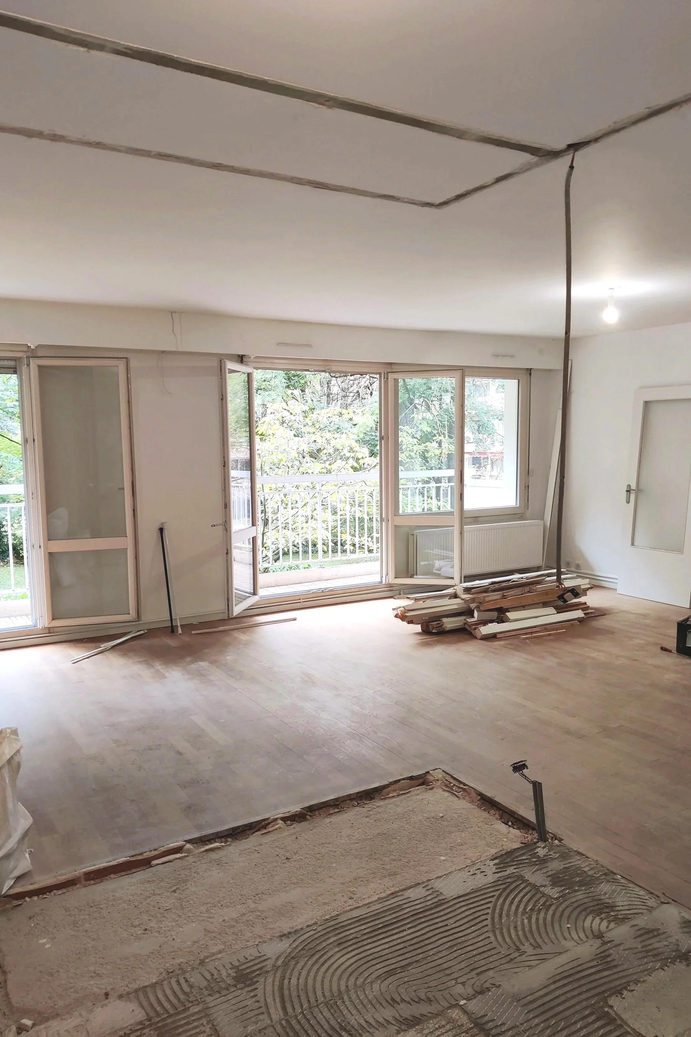 Salon en cours de rénovation avec parquet en bois, grande fenêtre avec porte-fenêtre ouvrant sur un balcon, piles de planches de bois, espace de travail en construction.