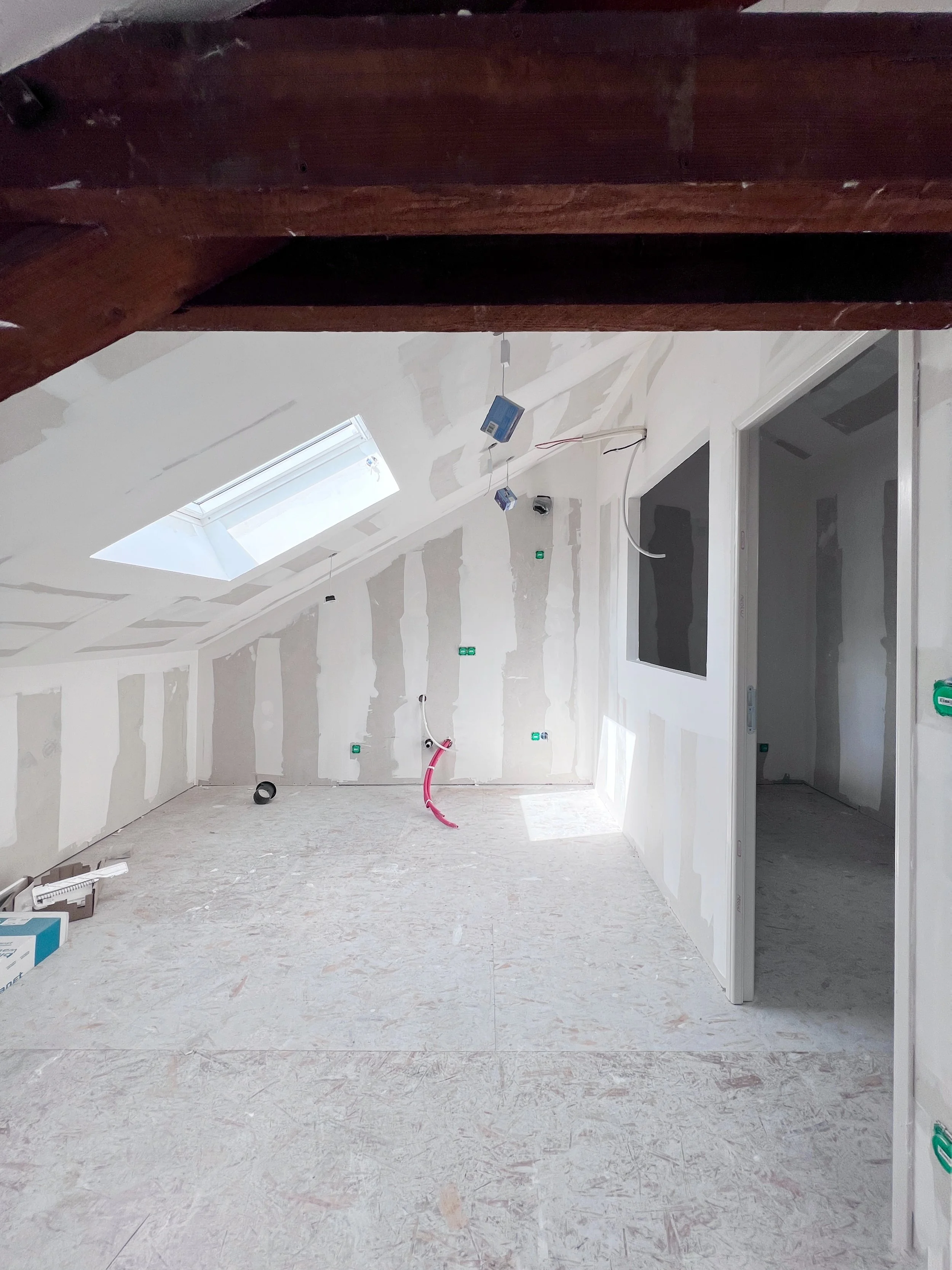Une pièce en cours de construction ou de rénovation, avec des murs partiellement peints ou recouverts d'enduit, un plafond avec un puits de lumière, et des câbles électriques suspendus. Le sol en sous-sol ou étage supérieur est en matériau brut, sans revêtement final.