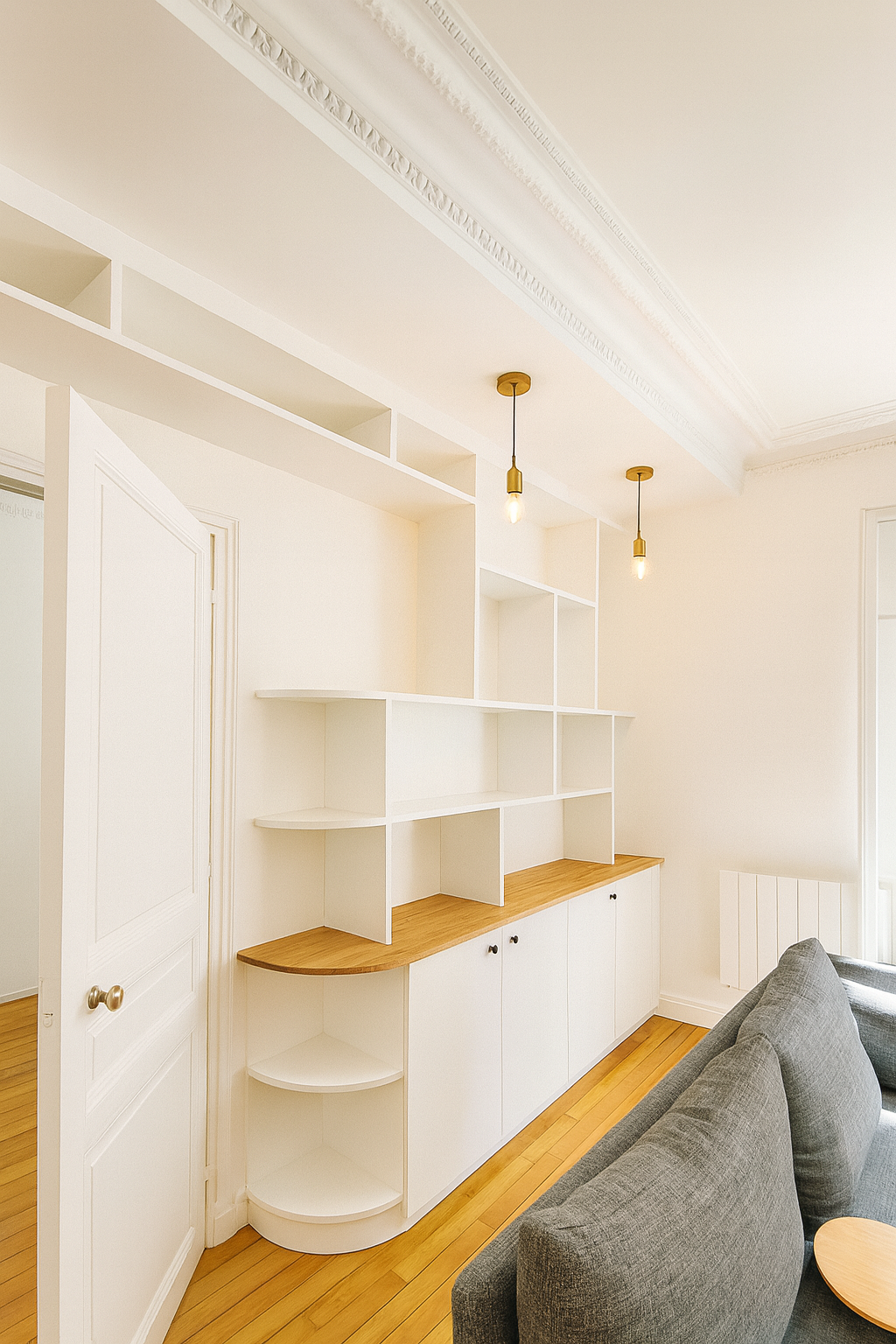 Intérieur d’un salon avec un mur blanc, un meuble en bois et blanc avec des étagères, deux lampes suspendues et un canapé gris.